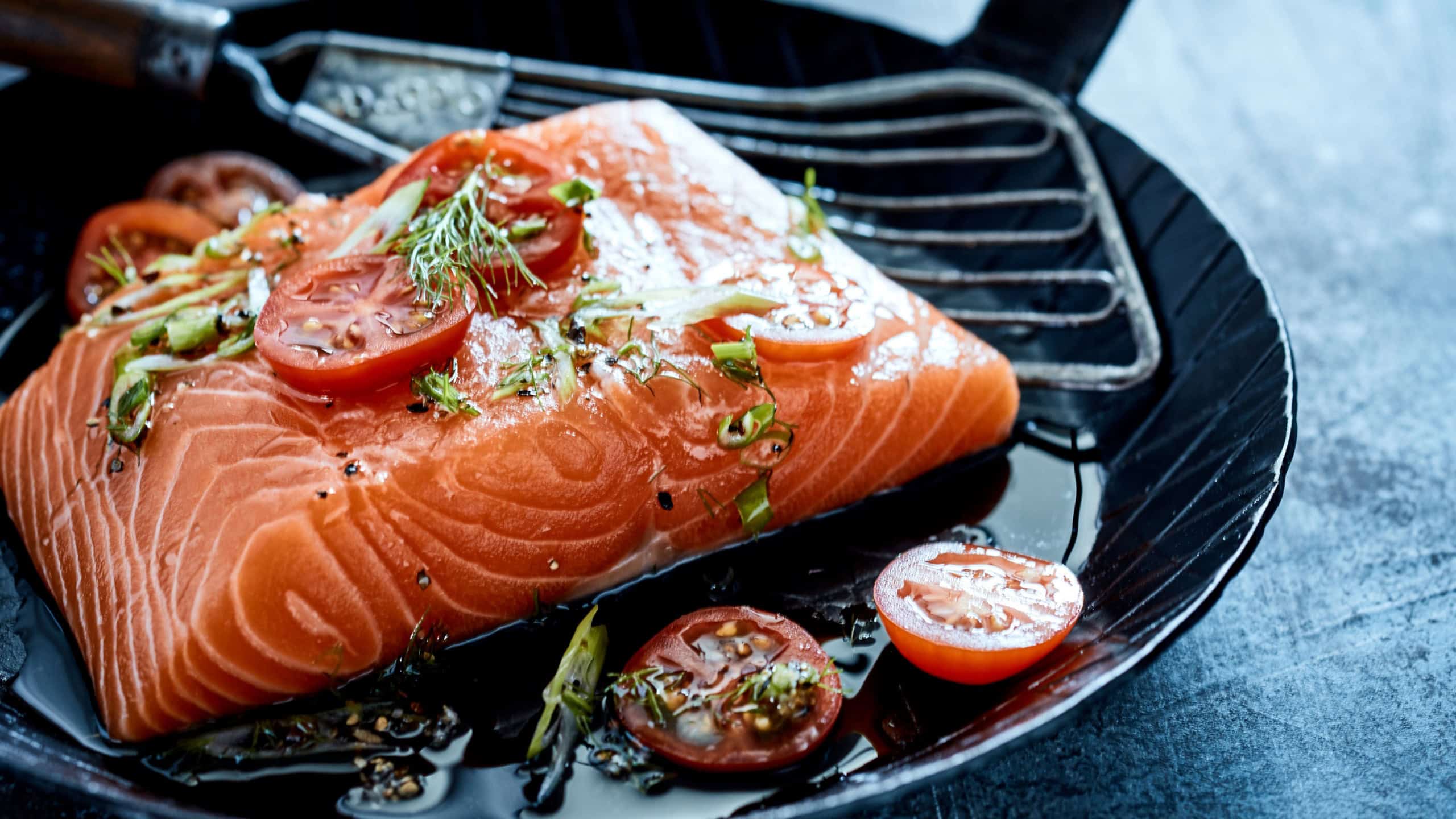 Fresh filleted salmon steak topped with chopped herbs and tomato on a small skillet with oil and spatula ready to be sauteed