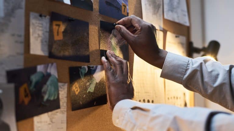 Hands pinning photos and notes on bulletin board in dimly lit room, with various pieces of evidence related to crime scene under investigation