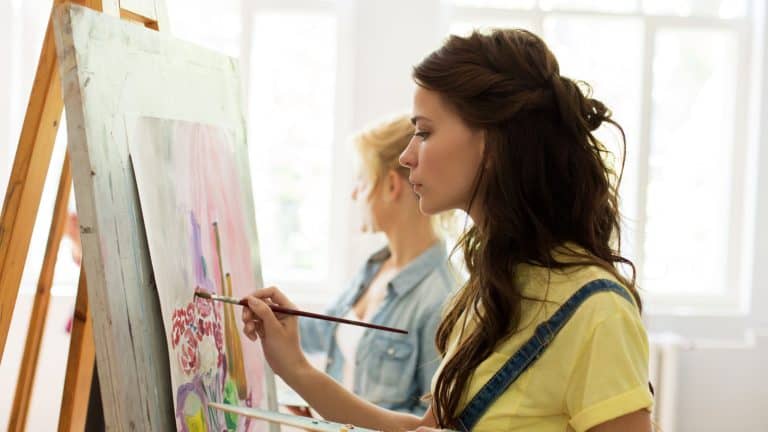 art school, creativity and people concept - woman with easel, palette and brush painting at studio