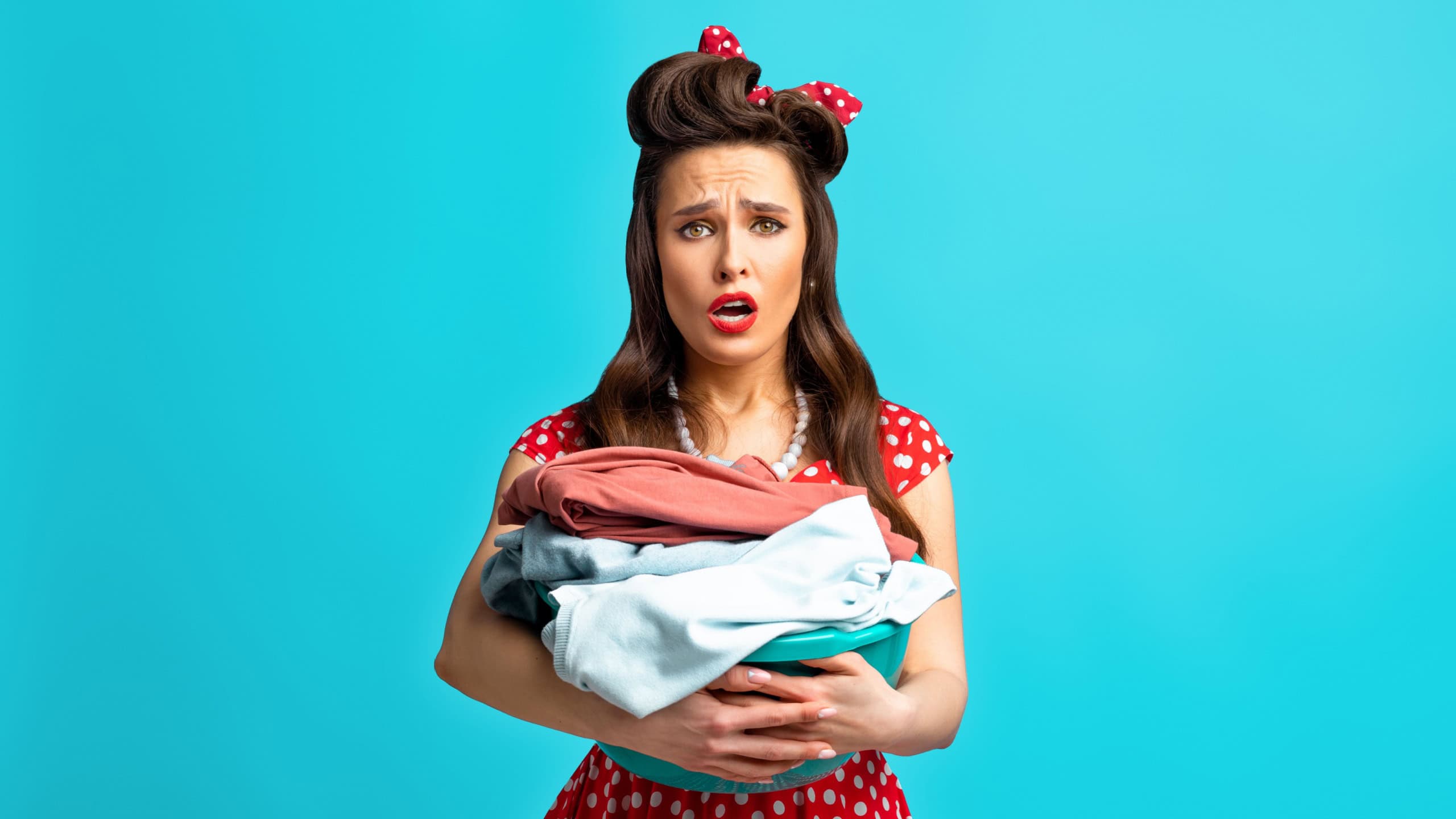 Unhappy tired young pinup woman in retro dress holding basin with laundry for washing or ironing on blue studio background. Classic style housewife sick of domestic duties, upset about house chores