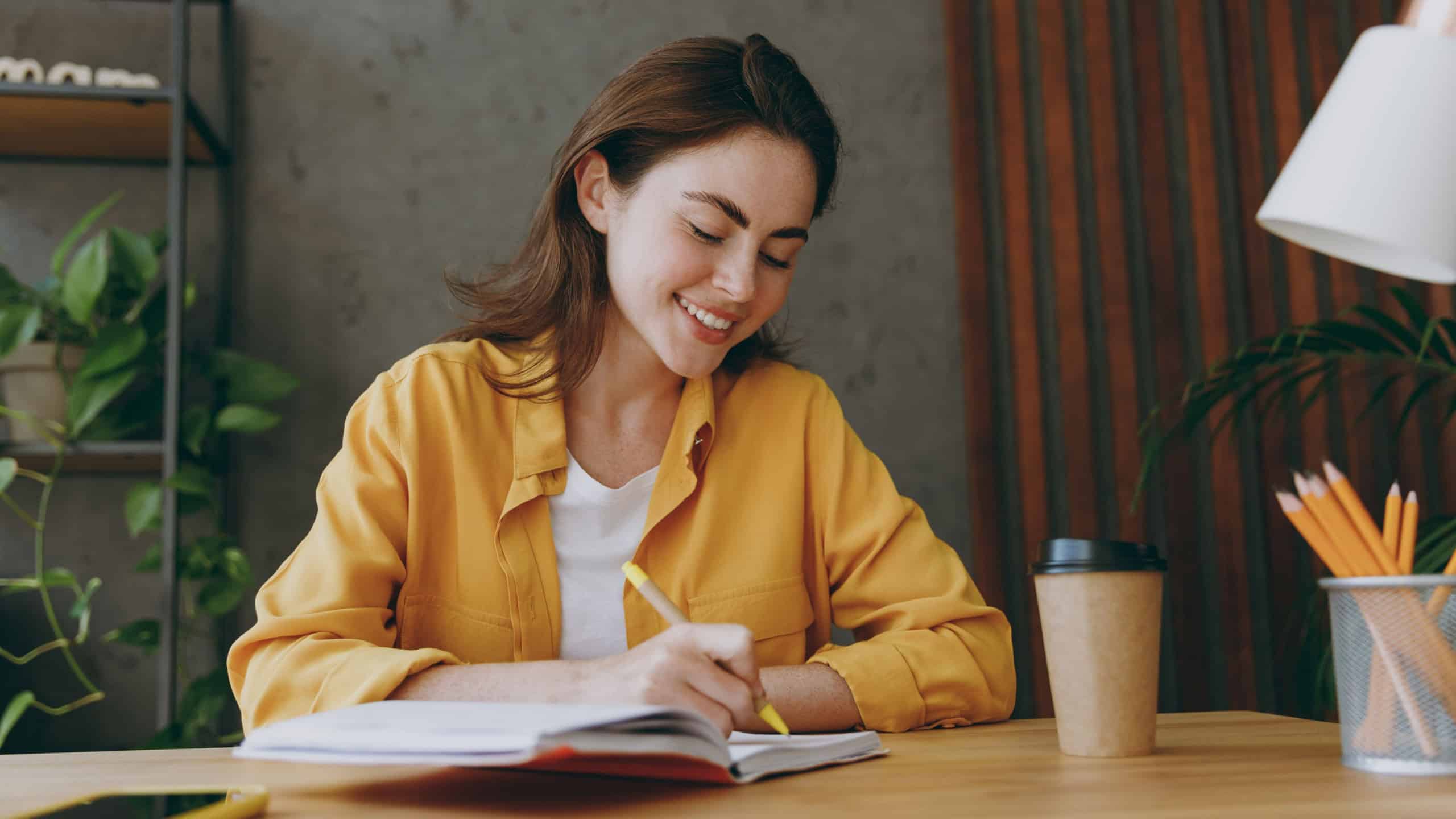 Young smiling happy cheerful fun european successful employee business woman 20s she wearing casual yellow shirt writing in notebook diary sit work at wooden office desk. Achievement career concept