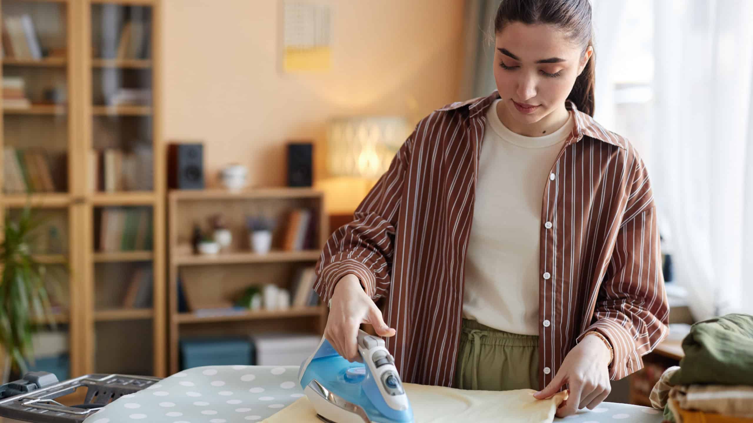 Waist up portrait of Caucasian young woman ironing clothes on ironing board while taking care of house chores on weekend copy space