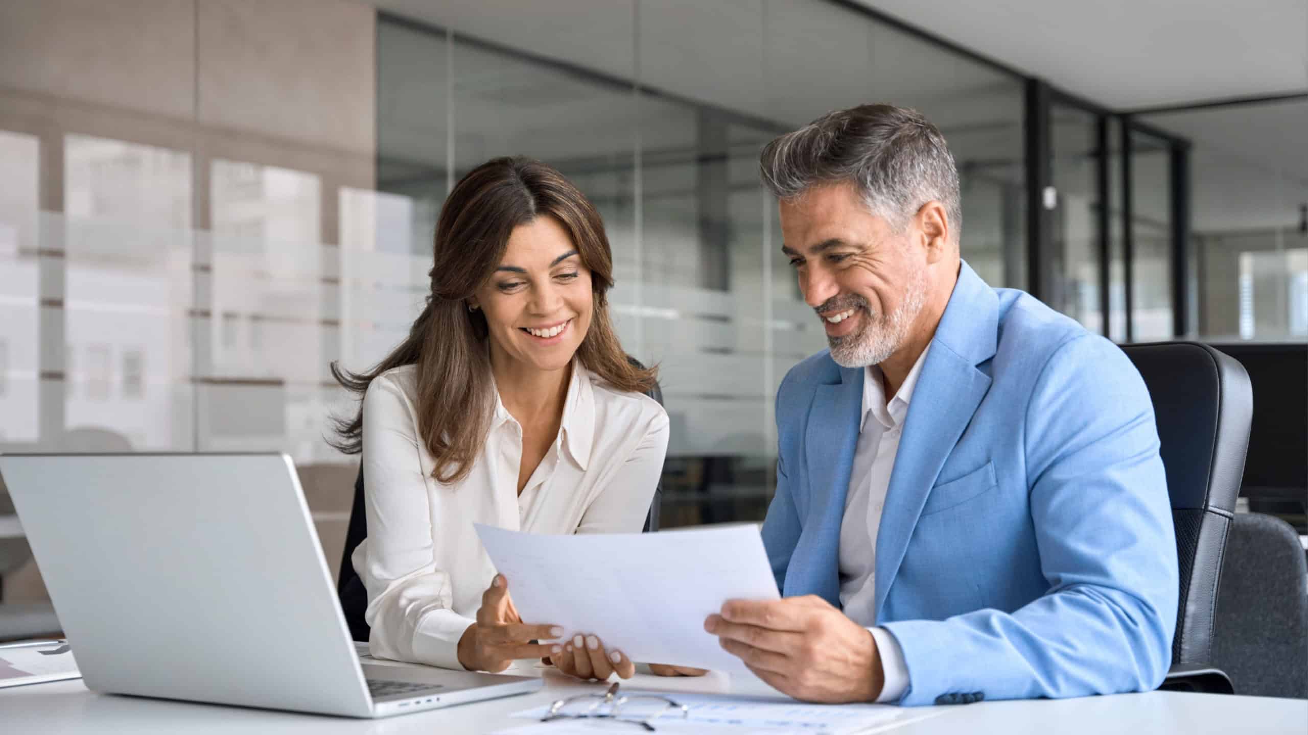 Two happy busy middle aged professionals man and woman business leaders partners checking document reading financial report talking working together on laptop computer in office at corporate meeting.