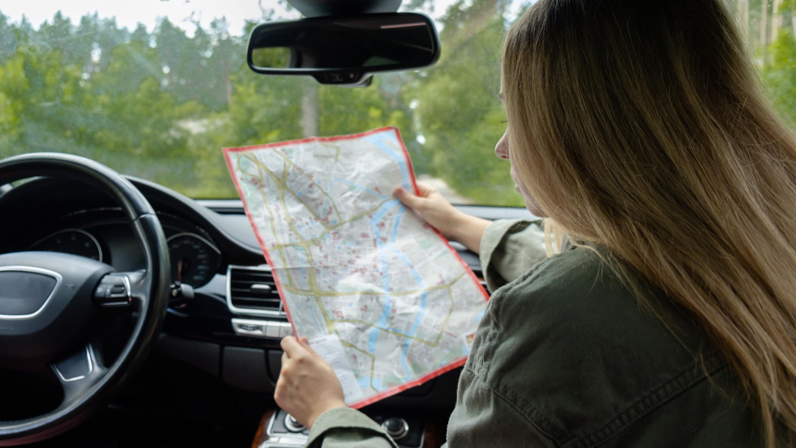 Blonde woman sitting inside the car checking the route on map to get to the destination. Afraid to get lost in forest. Young tourist explore local travel making candid real moments. True emotions