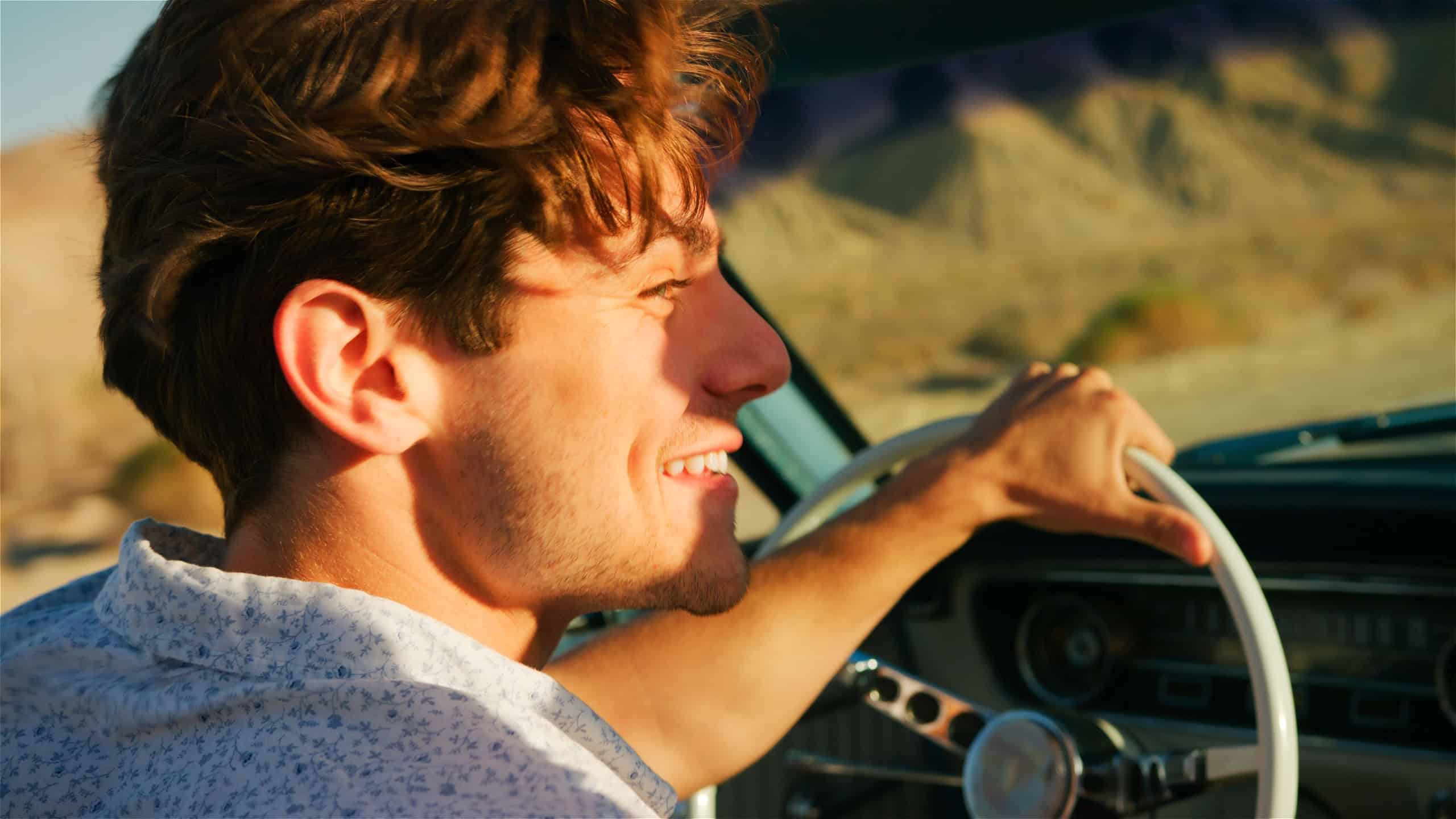 Close Up Of Man Driving Classic Open Top Car On Road Trip Through Countryside At Sunset
