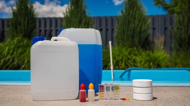 Swimming pool cleaning setup with chlorine tablets, liquid chemical canisters, and pH test kit on concrete near water under bright sunlight. High quality photo