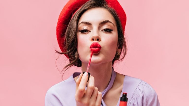 Portrait of young girl in red beret painting her lips with bright lipstick on pink background
