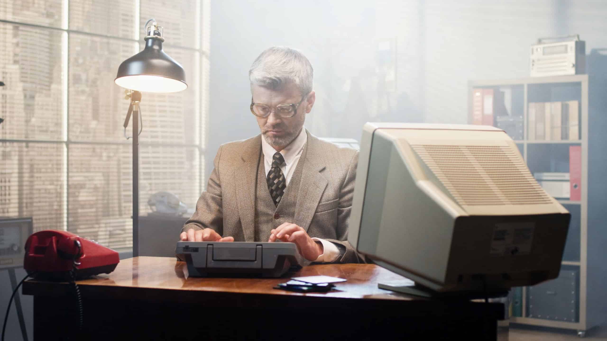 Retro caucasian male working in vintage interior of office. Man in glasses sitting at old computer and examining diskettes at the table. Serious businessman of 80's