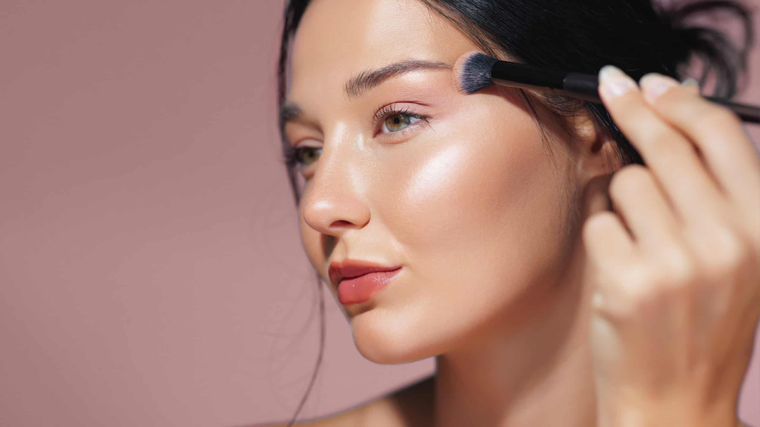 Detailed close-up of a young woman applying shimmering highlighter on her cheekbone with a makeup brush, highlighting her radiant and flawless skin. Ideal for beauty, makeup, and skincare promotions.