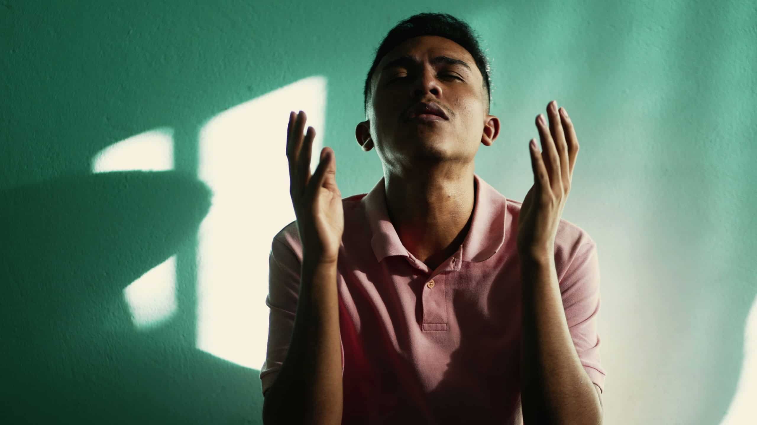 Spiritual young hispanic man praying to God at home. Faithful believer South American male person looking at sky pleading grace