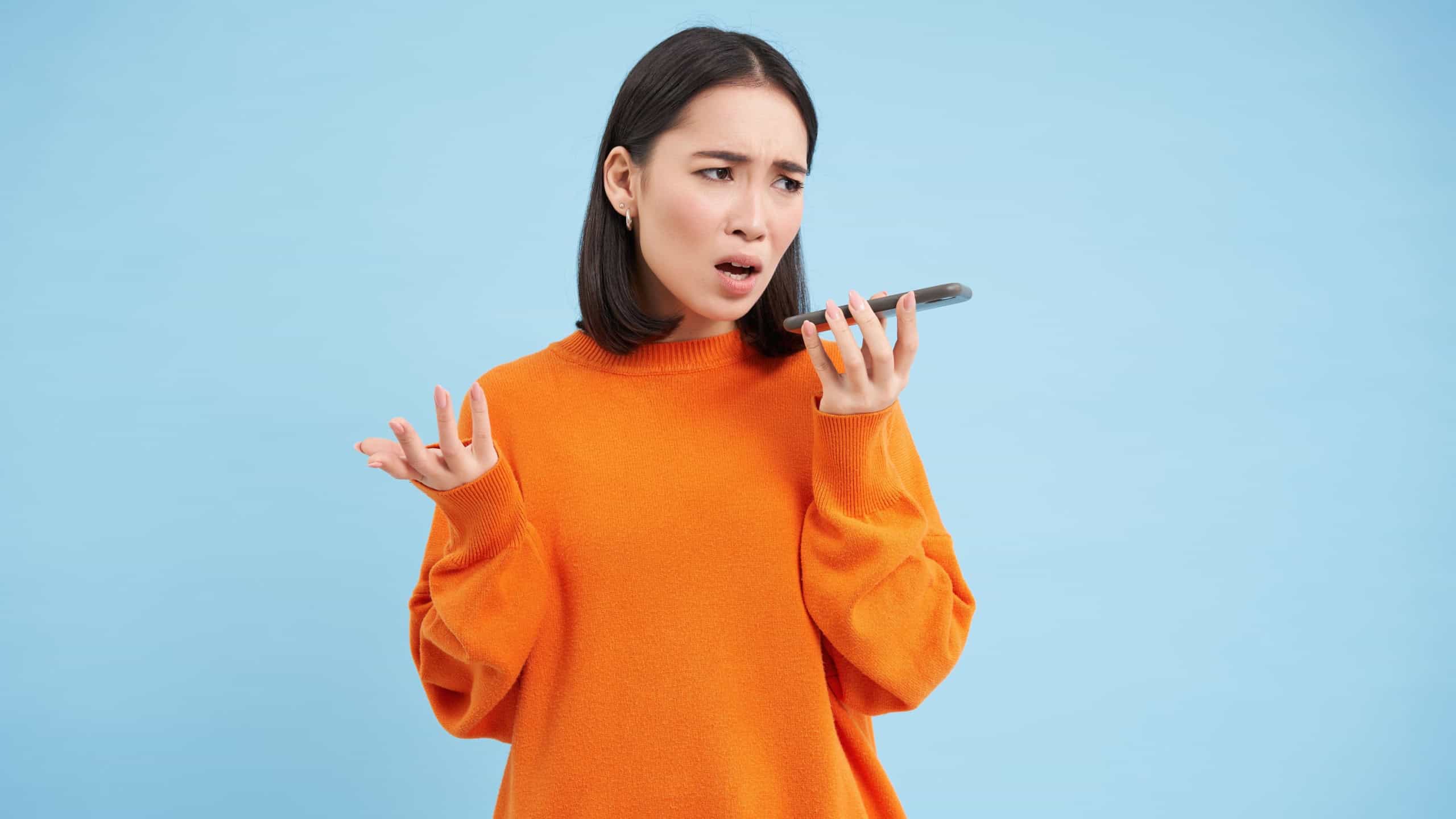 Portrait of upset young woman records voice mail or message, complains with disappointed, sad face, standing over blue background and holding mobile phone.