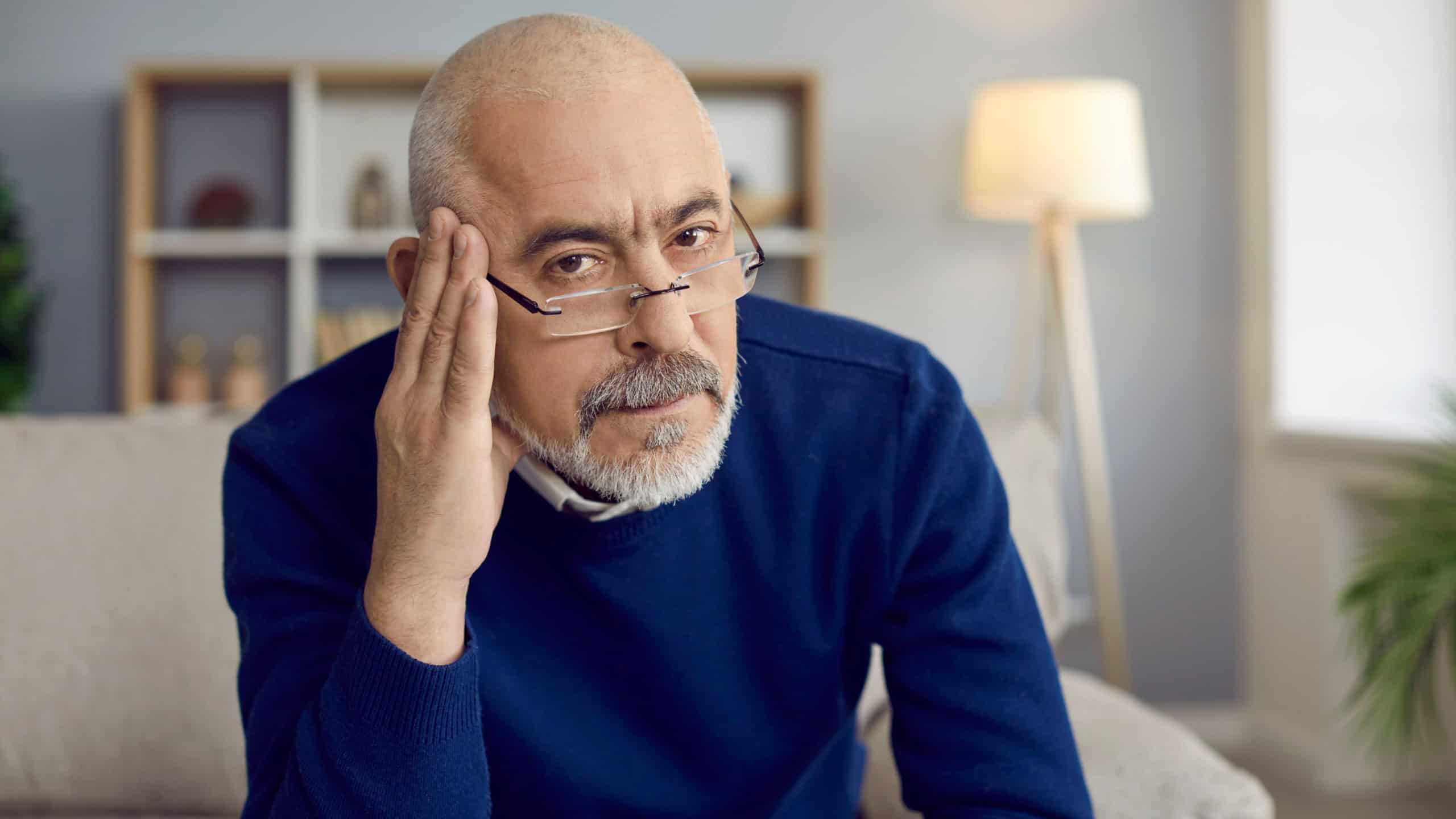 Portrait of strict reserved senior man in glasses sitting on couch with serious face, feeling skeptic, doubtful, uninspired and unexcited about bad idea or having secondhand embarrassment for somebody
