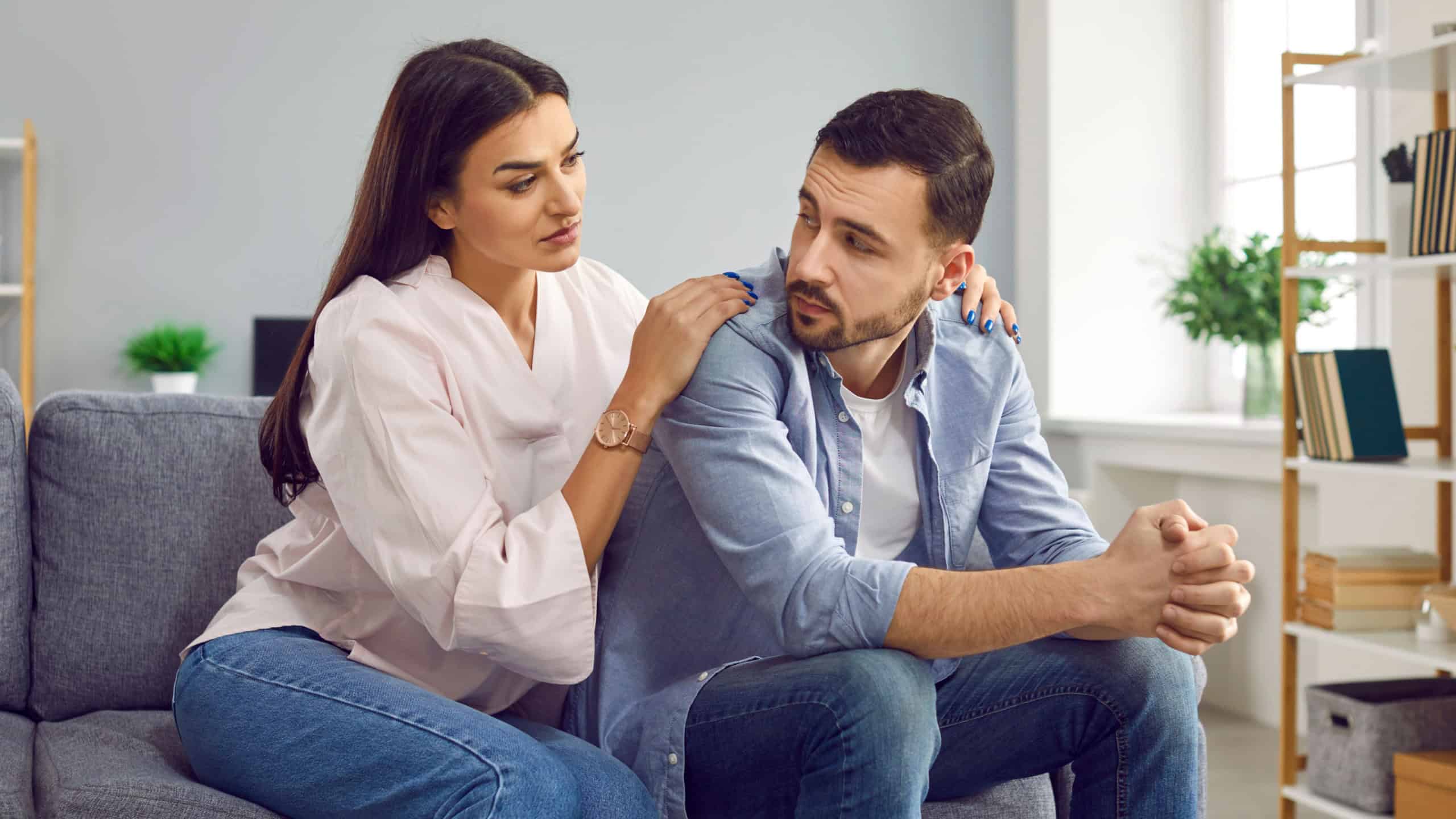 Attractive wife hugging husband from back to comfort or apologize. Young woman hugging upset man, expressing understanding, saying sorry, showing support. Couple sitting on sofa at home