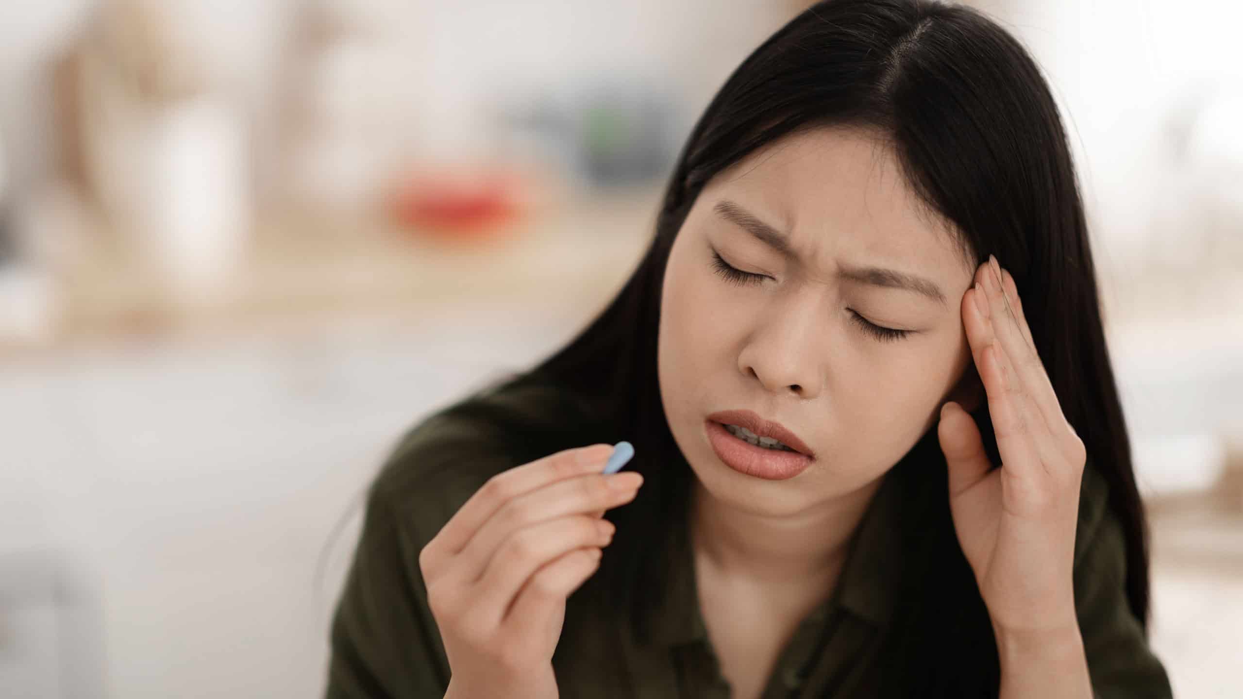 Closeup of unhappy distressed young asian woman sit at kitchen desk with closed eyes, touch her head, holding pill in her hand. Sad chinese lady taking painkiller, suffer from migraine, copy space