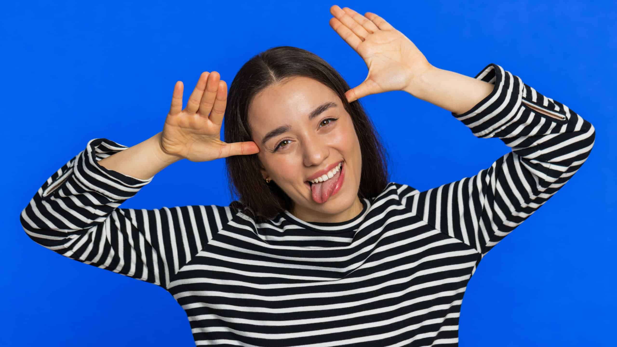 Funny Caucasian woman showing tongue making faces at camera, fooling around, joking, aping with silly face, teasing, bullying, abuse disrespect. Joyful young girl isolated on blue background indoors