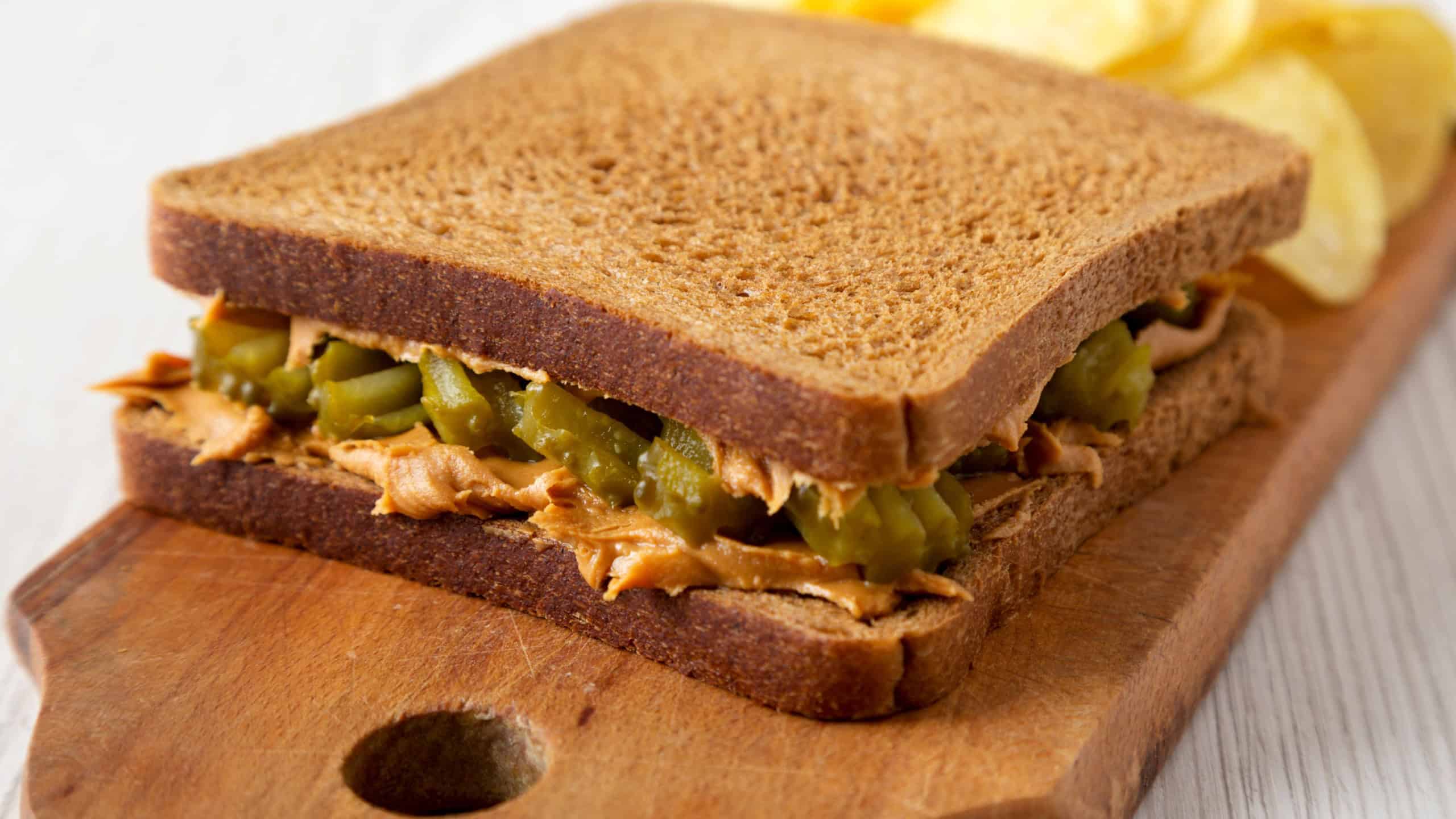Homemade Peanut Butter Pickle Sandwich with Potato Chips on a rustic wooden board on a white wooden surface, side view. Close-up.