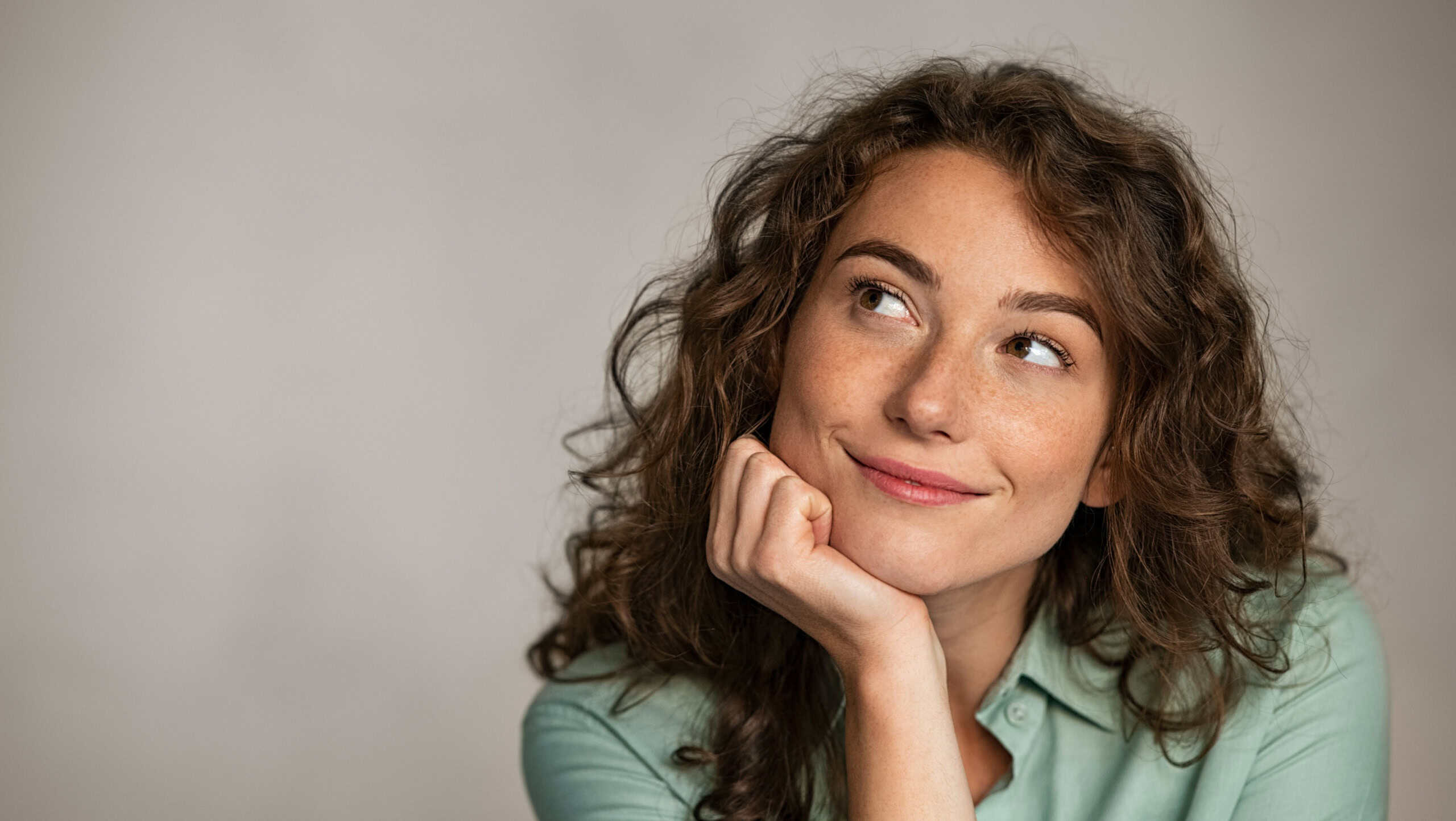 Portrait of young thoughtful woman with hand on chin having an idea against grey background. Pensive woman looking away while thinking. Close up face of girl planning her future isolated on gray wall.
