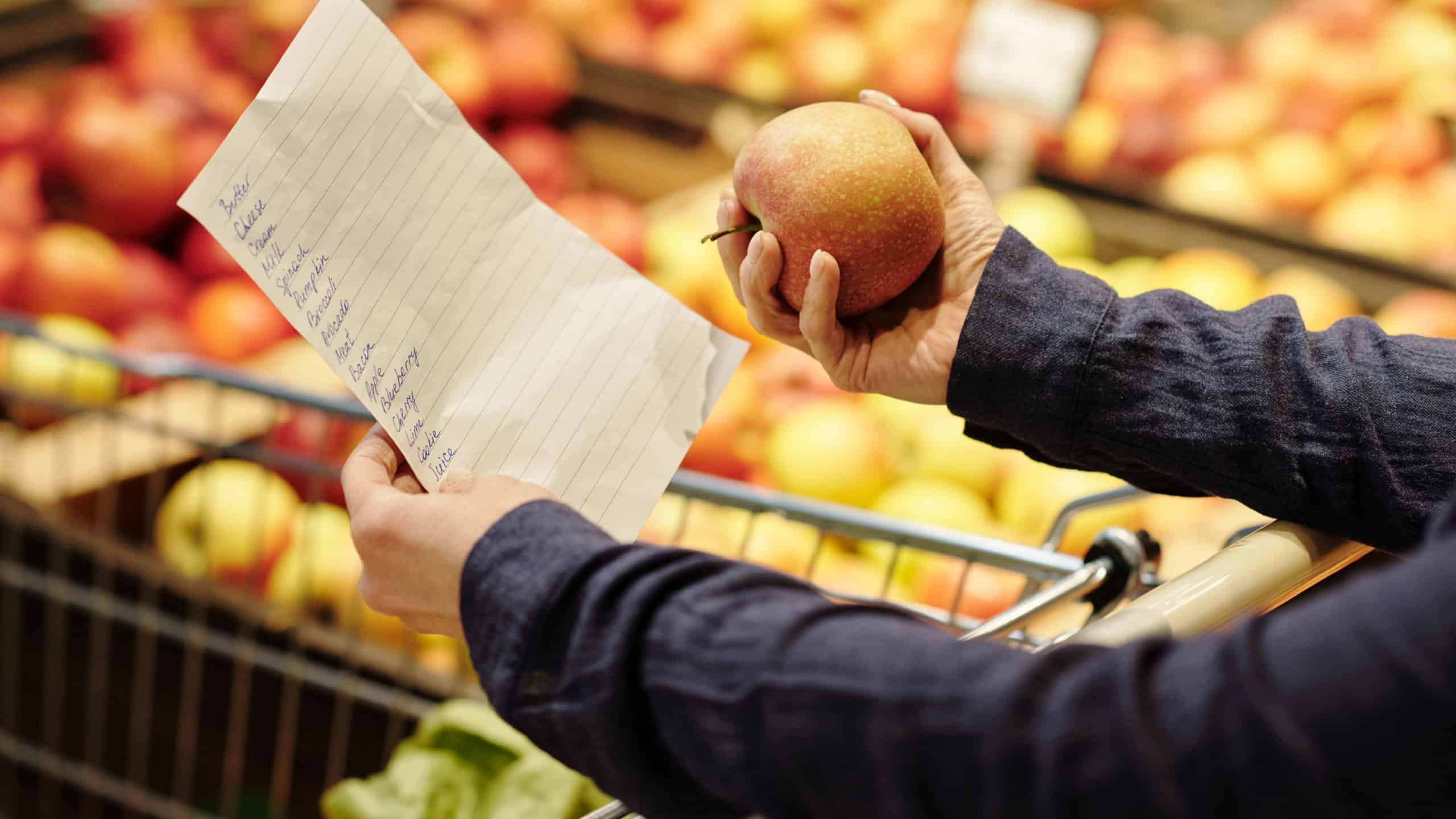 Close-up of mature female customer hands holding shopping list and fresh ripe red apple while choosing fruits in grocery department