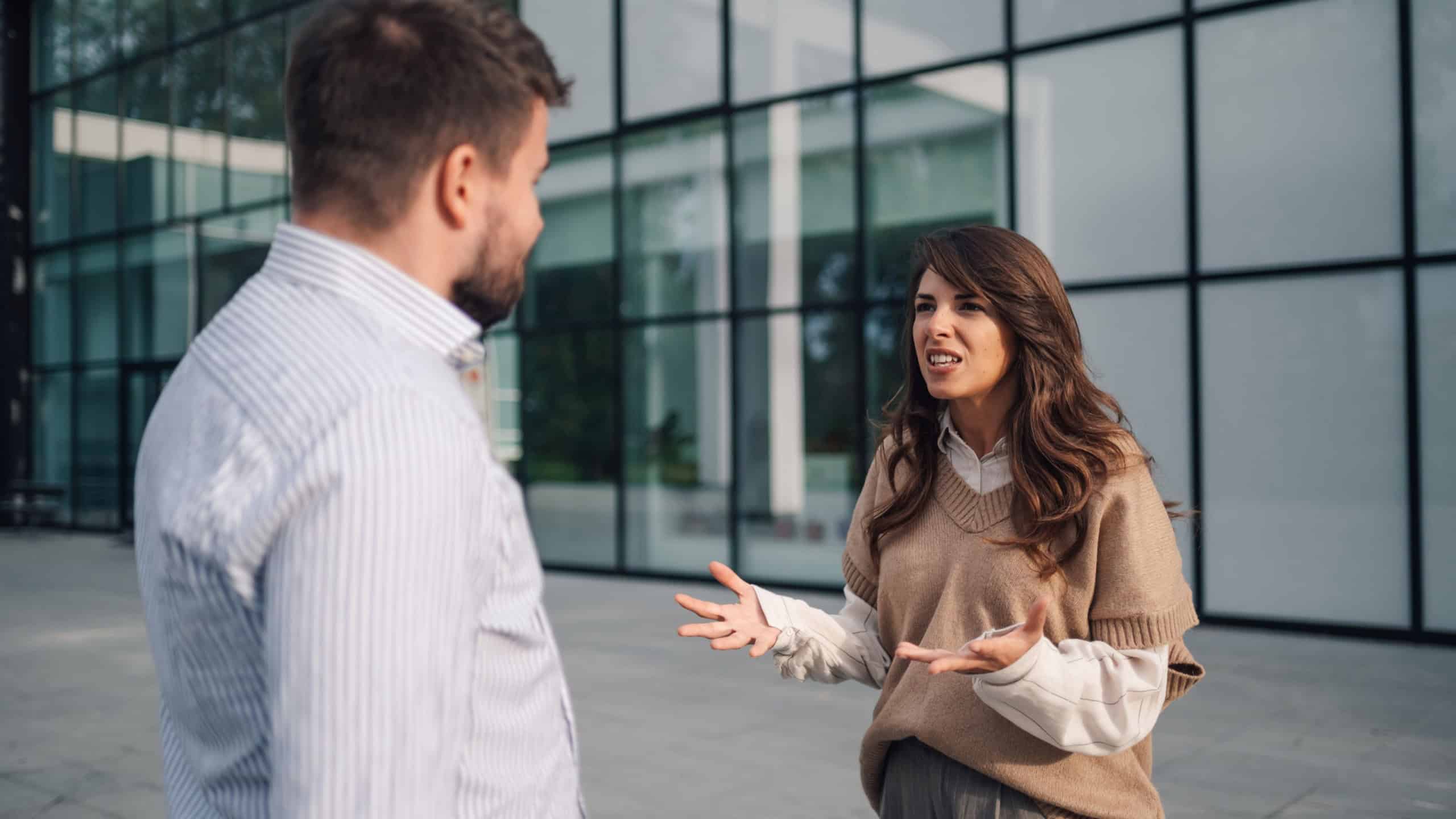 Two colleagues arguing in front of a modern office building in the city, showing frustration and disagreement in a corporate environment