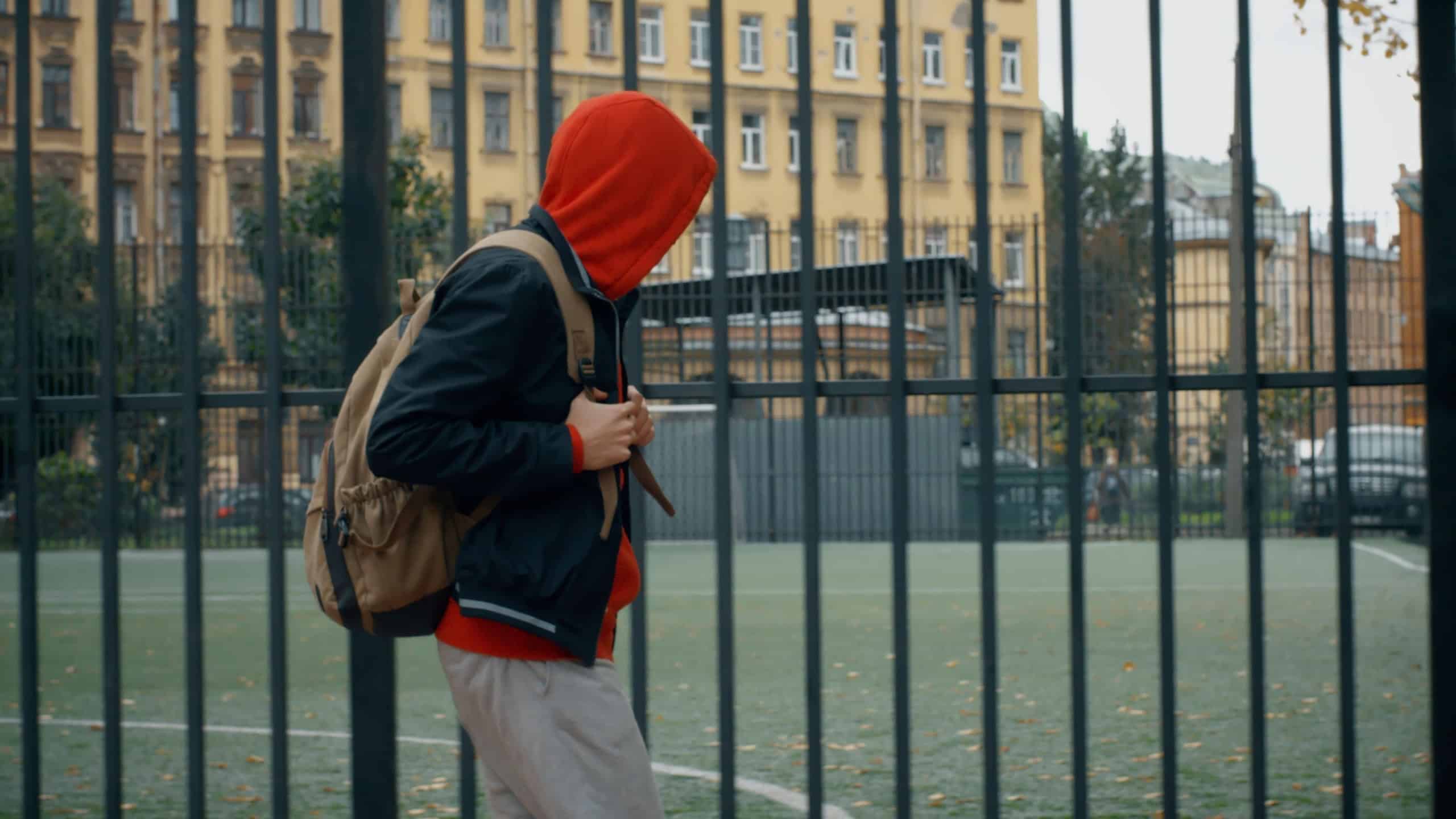 Teenage boy with backback walking near school football field. Side view of teenager walk to school playground in city