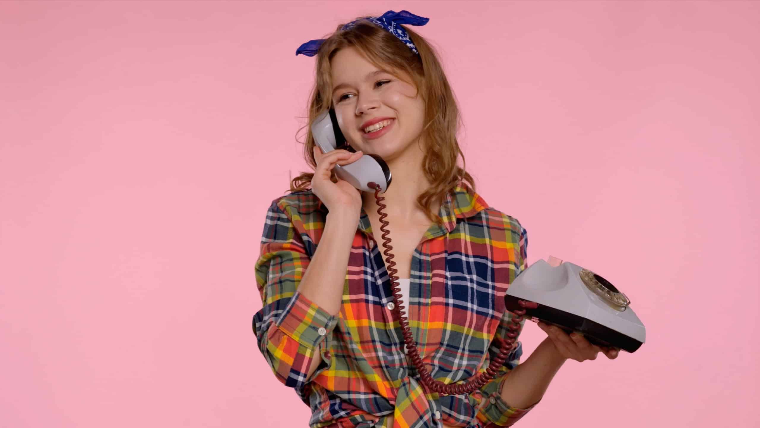 Smiling girl in a plaid shirt and bandana talks on a vintage rotary phone against a pink background.