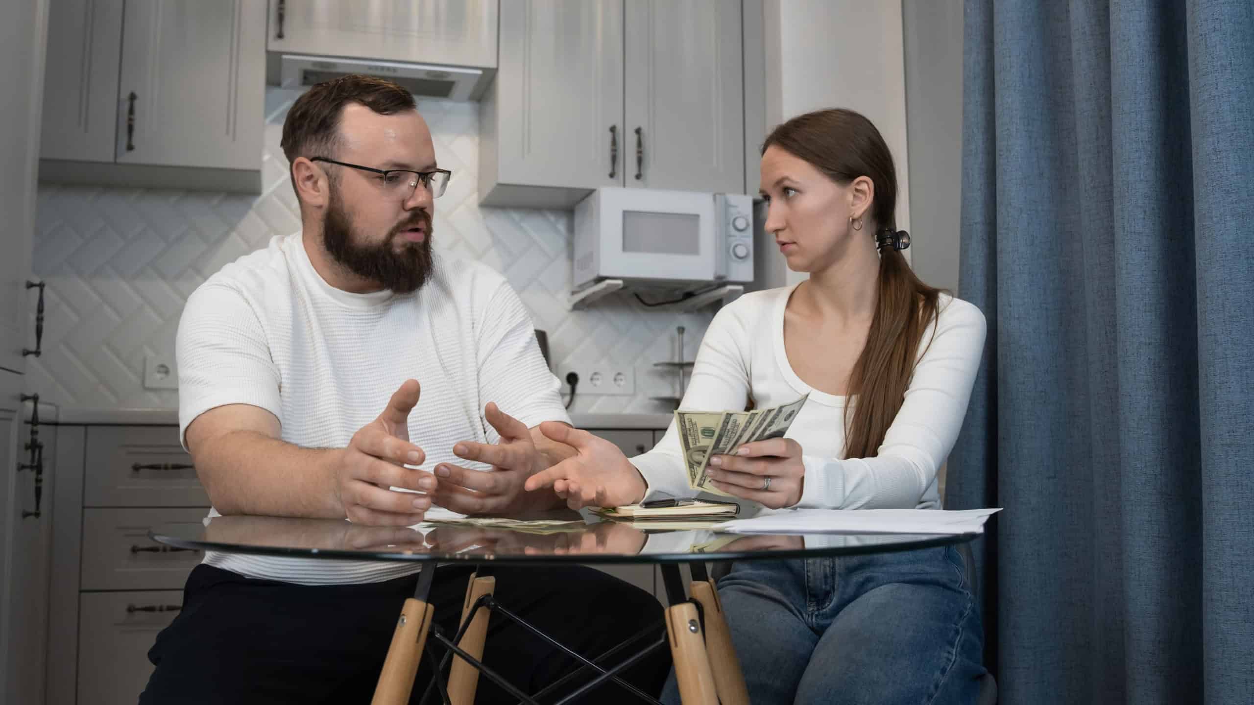 Couple sitting at a kitchen table, discussing finances and managing their household budget while holding money, representing financial stress and planning