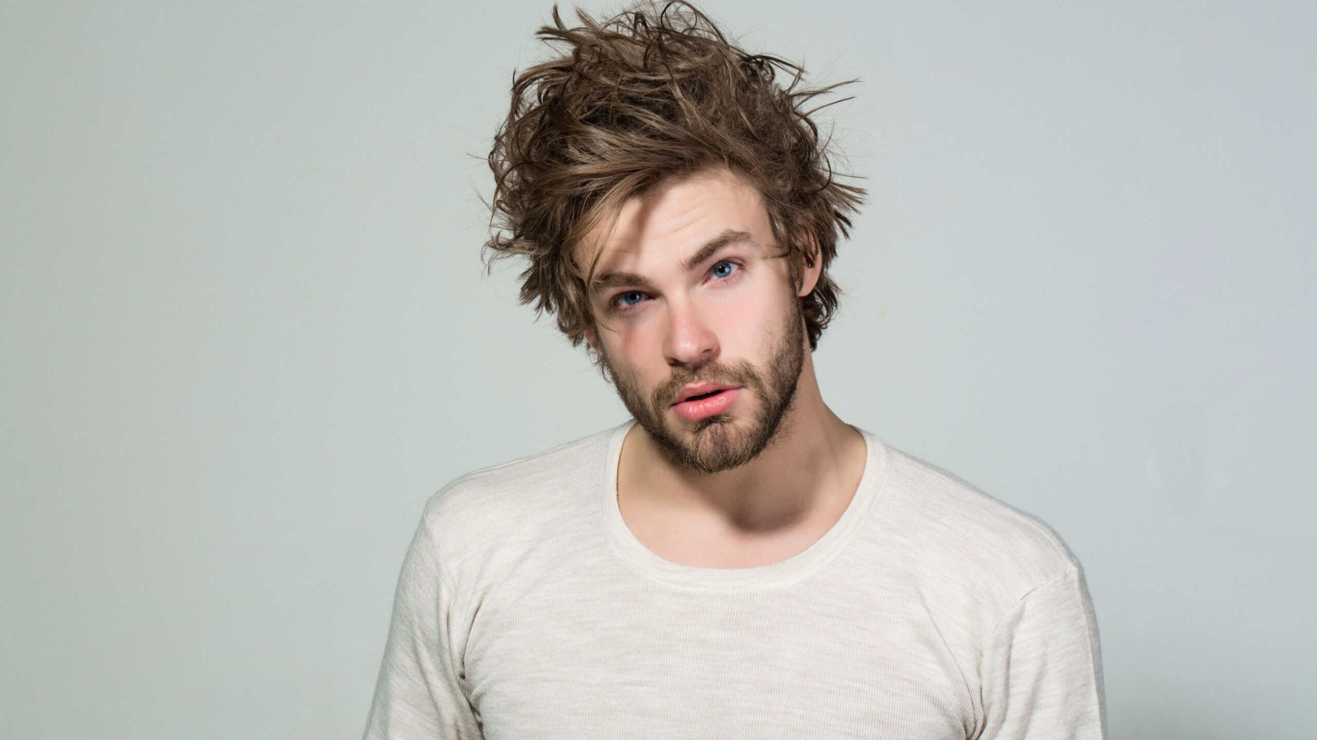 sleepy man with disheveled and uncombed long hair with beard on face in white underwear on grey background, morning and wake up, everyday life, barbershop