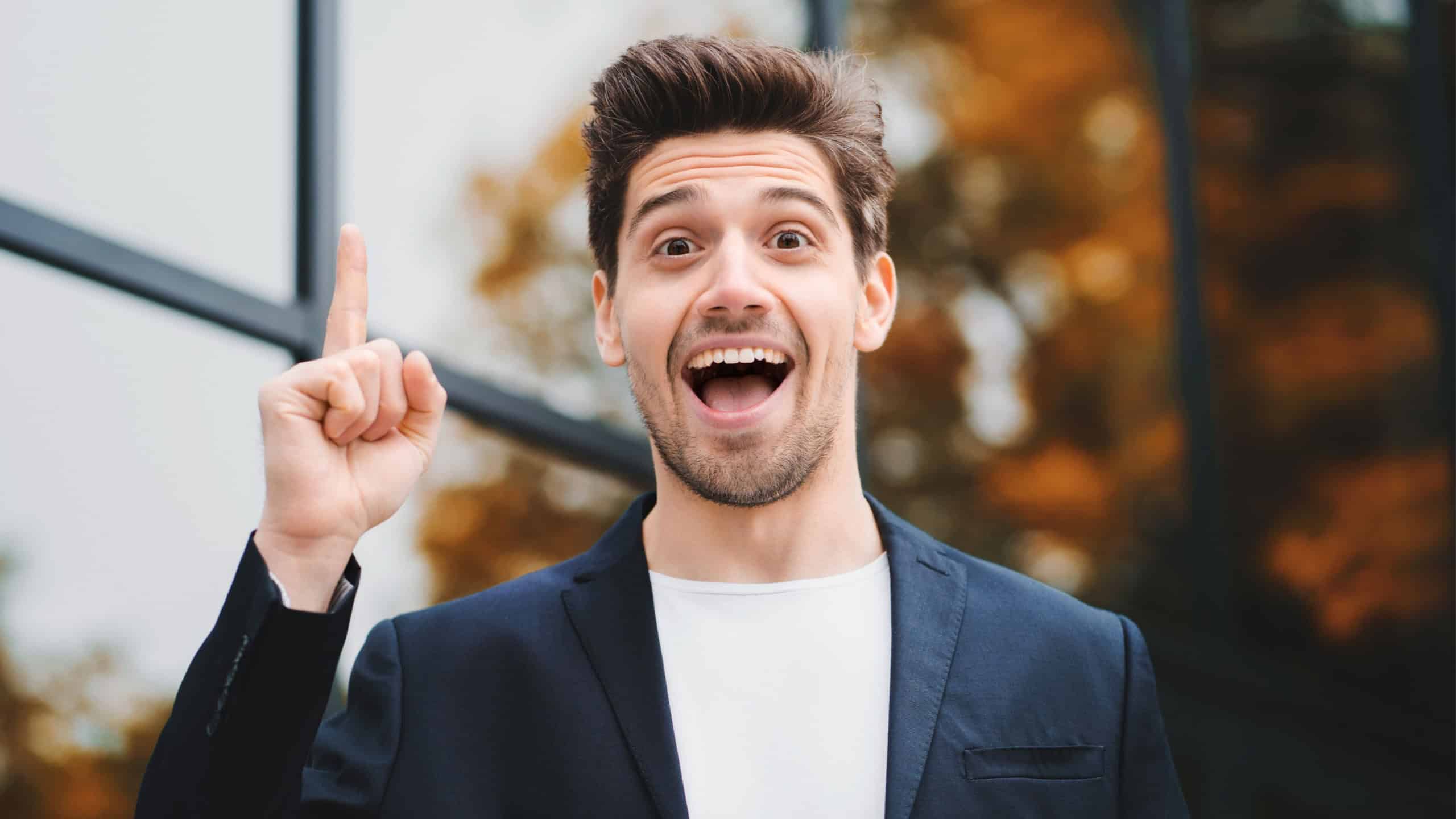 Portrait of young thinking pondering businessman having idea moment pointing finger up on office building background. Smiling happy student man showing eureka gesture.