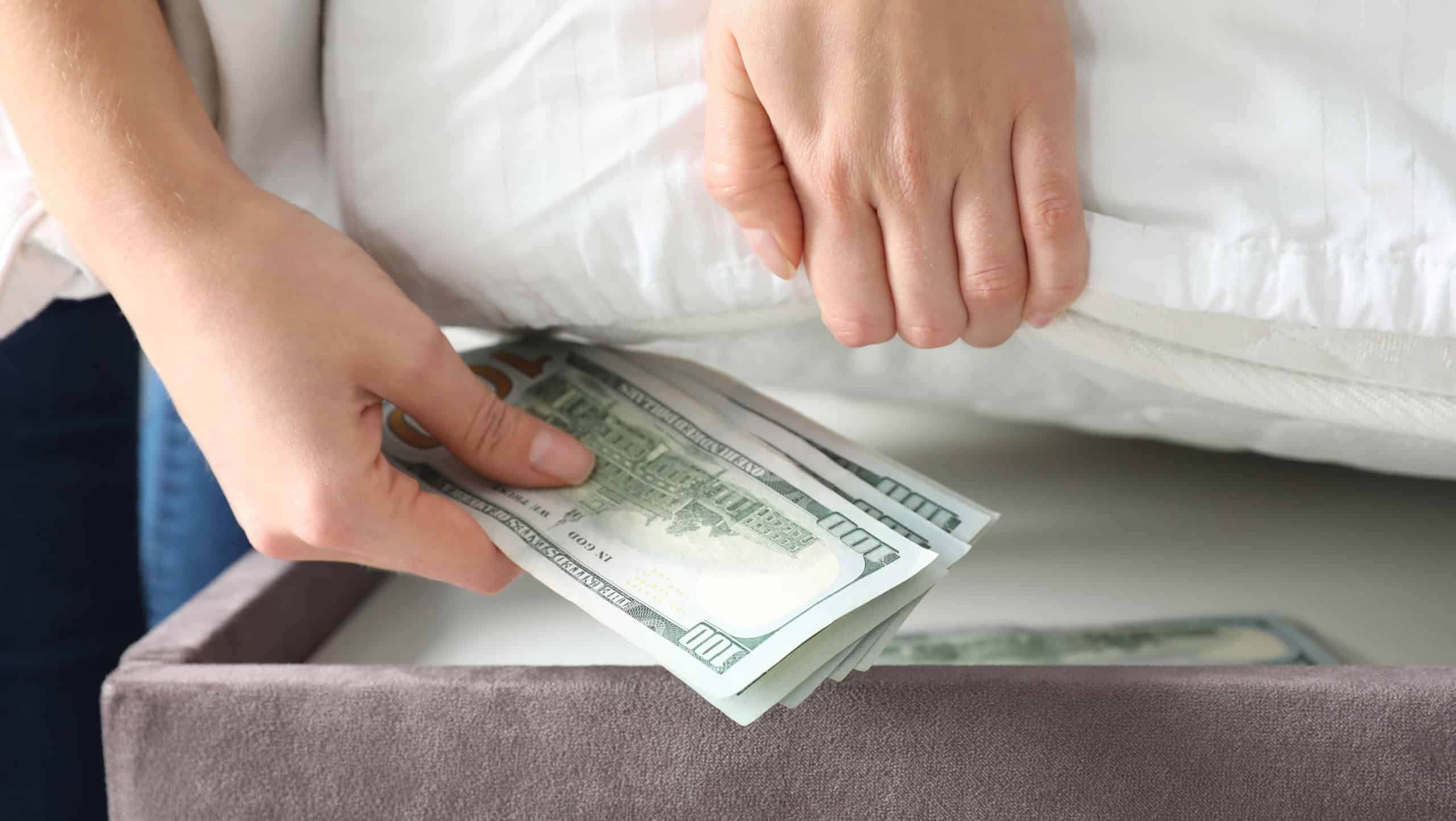 Woman hiding dollar banknotes under mattress in bedroom, closeup. Money savings