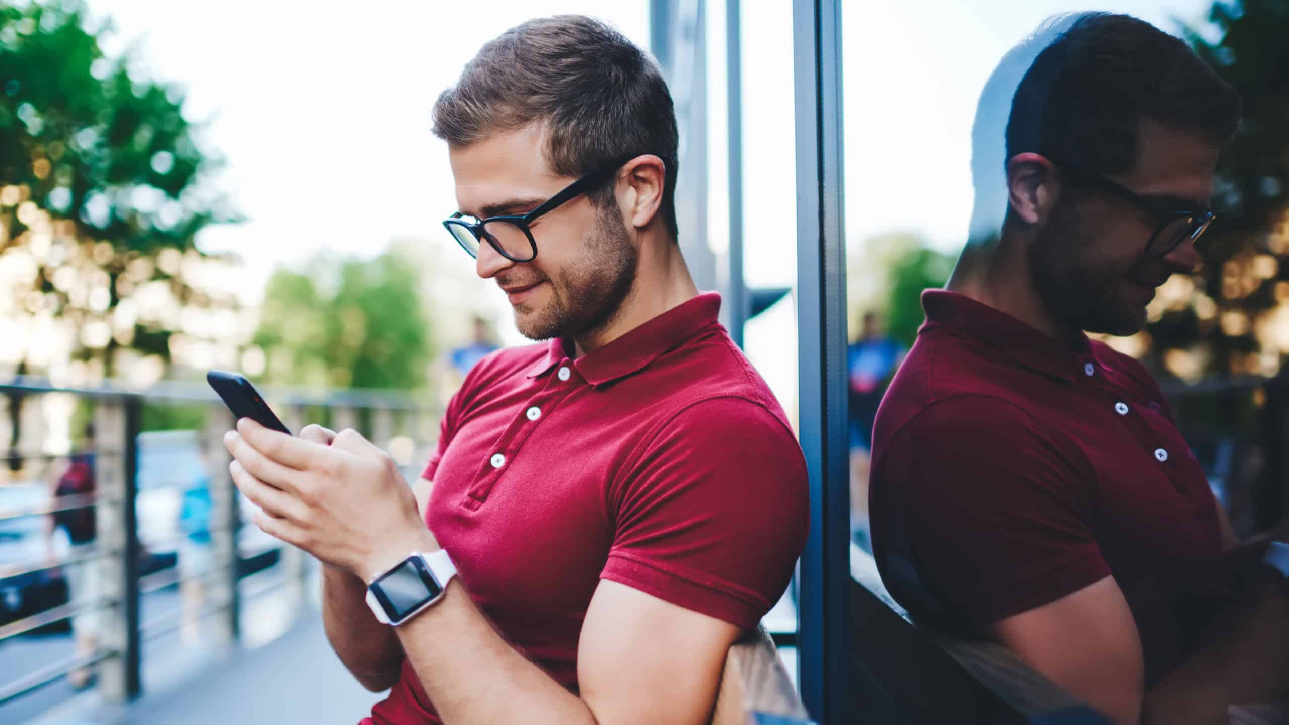Side view of happy young man wearing eyeglasses and smart watch sitting on bench on urban background and smiling while surfing social network in smartphone