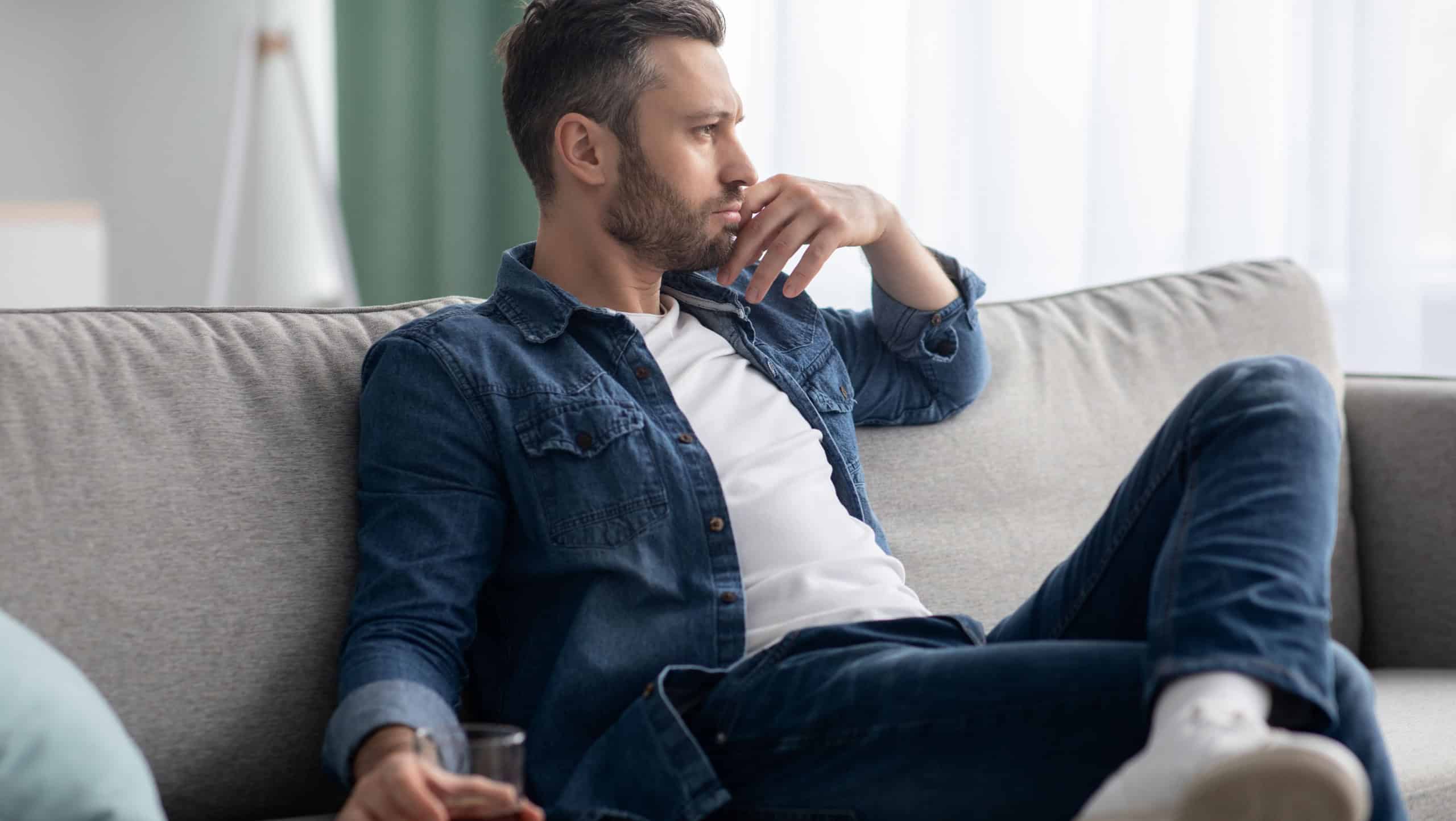 Depression, crisis concept. Sad middle-aged bearded man sitting on sofa, drinking alcohol at home, feeling lonely and sick, holding glass of whiskey and looking through the window, copy space
