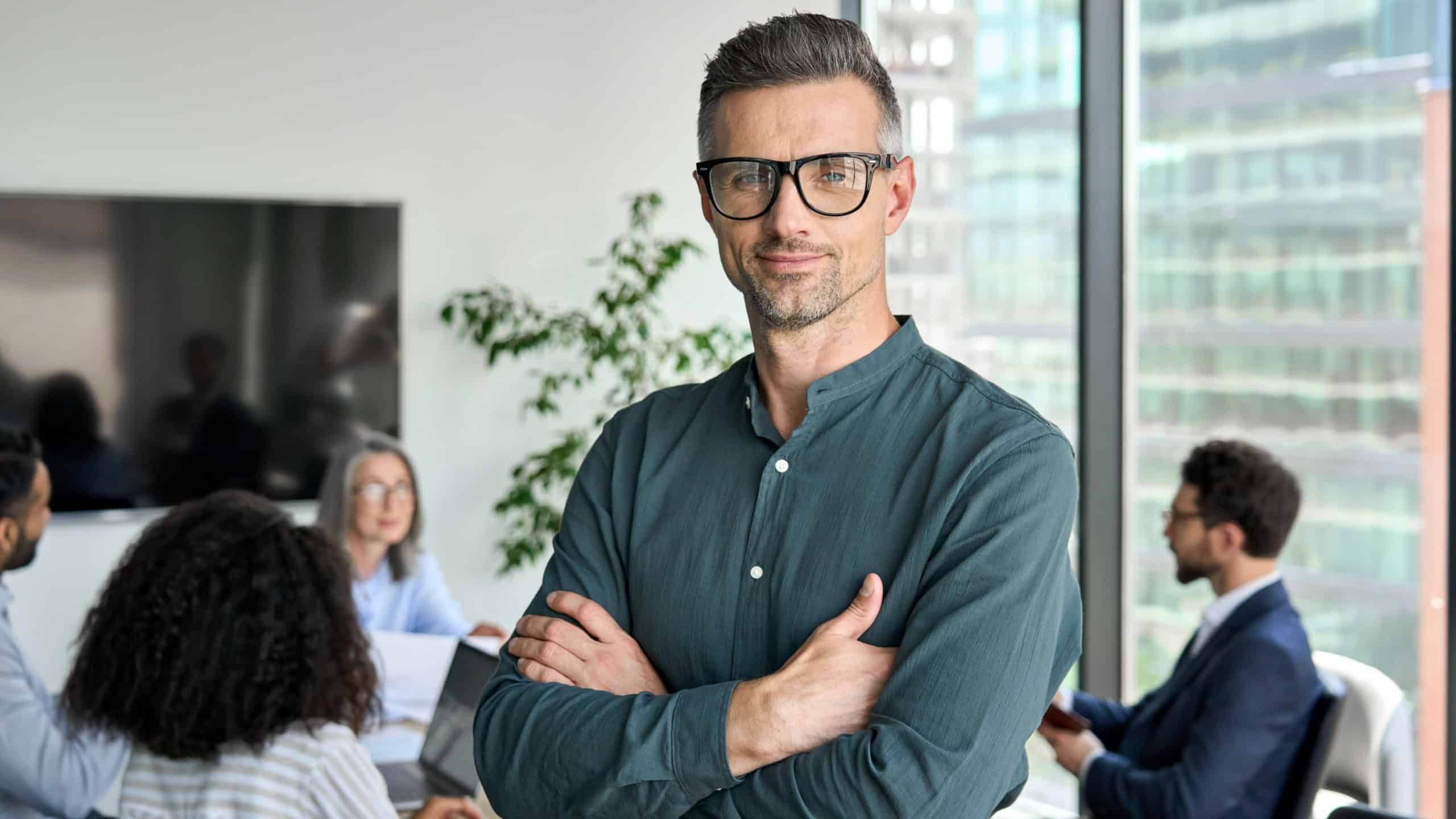 Smiling confident mature businessman leader looking at camera standing in office at team meeting. Male corporate leader ceo executive manager wearing glasses posing for business portrait arms folded.