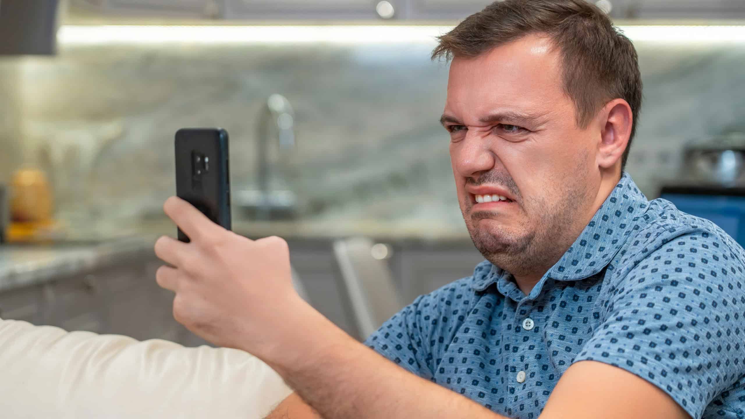 Annoyed millennial man with disgust look at smartphone screen at modern living room. Portrait of manager cringe and looking at phone screen with squeamishness, bad joke or inappropriate content.