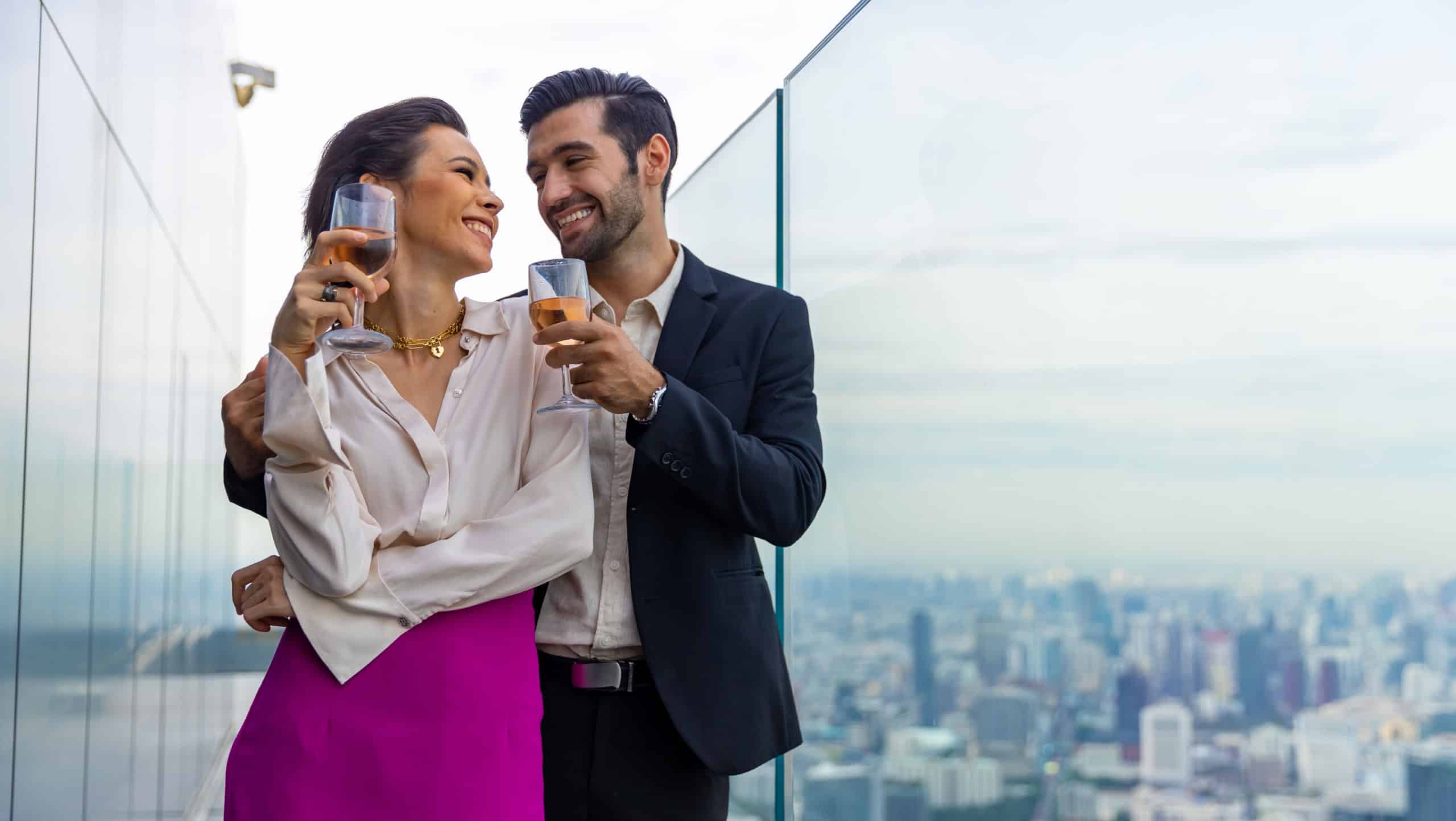 Caucasian couple celebrating at luxury skyscraper rooftop bar at summer sunset. Confident man and woman couple enjoy city lifestyle having dinner and cocktail drink at outdoor restaurant in evening.