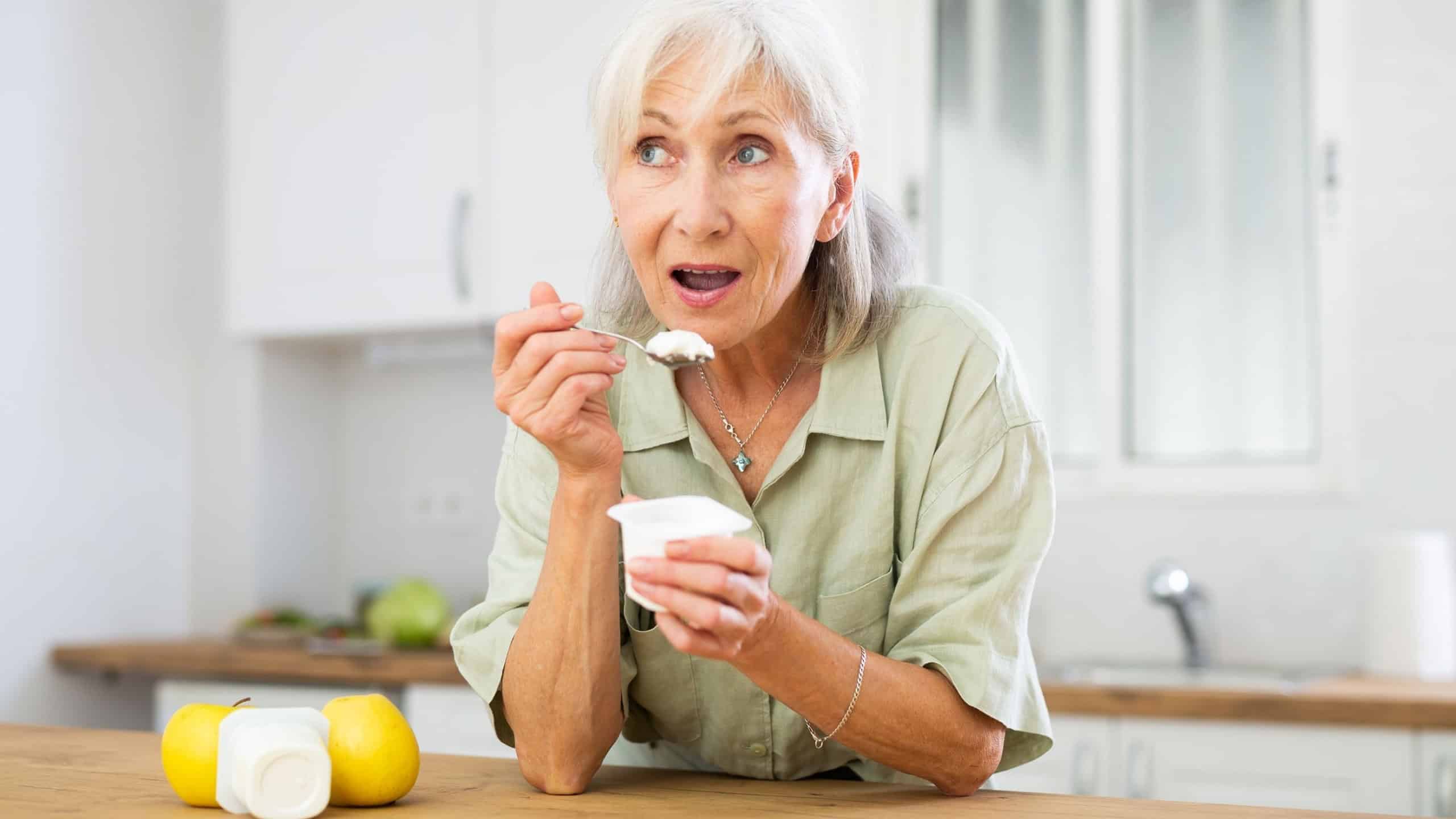 Senior woman eating yogurt in kitchen in apartment. Old woman enjoying meal, eating dairy product.