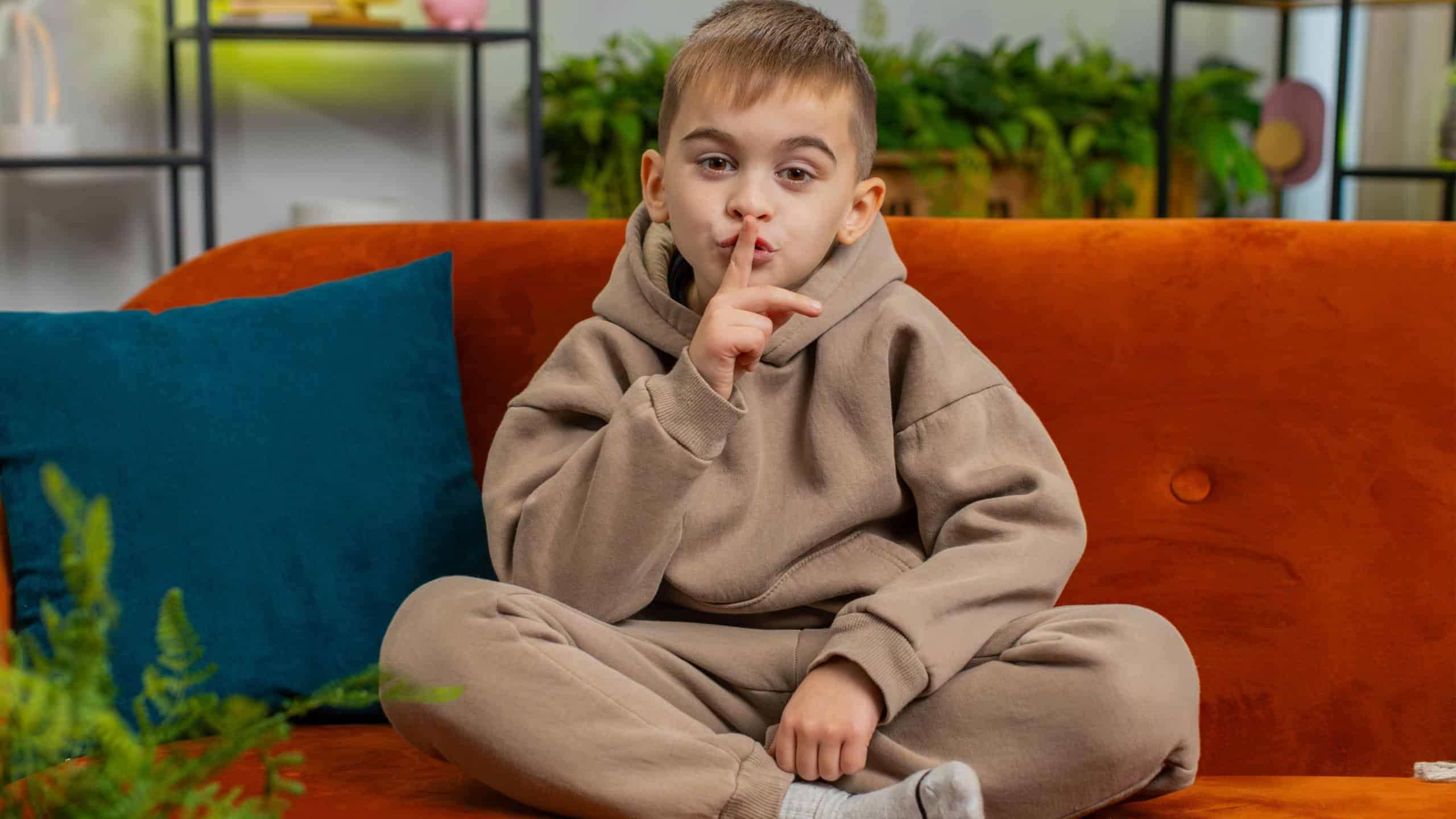 Shh be quiet please. Preteen child boy presses index finger to lips makes silence hush gesture sign do not tell gossip secret. Teenager male little kid at home apartment living room sits on couch