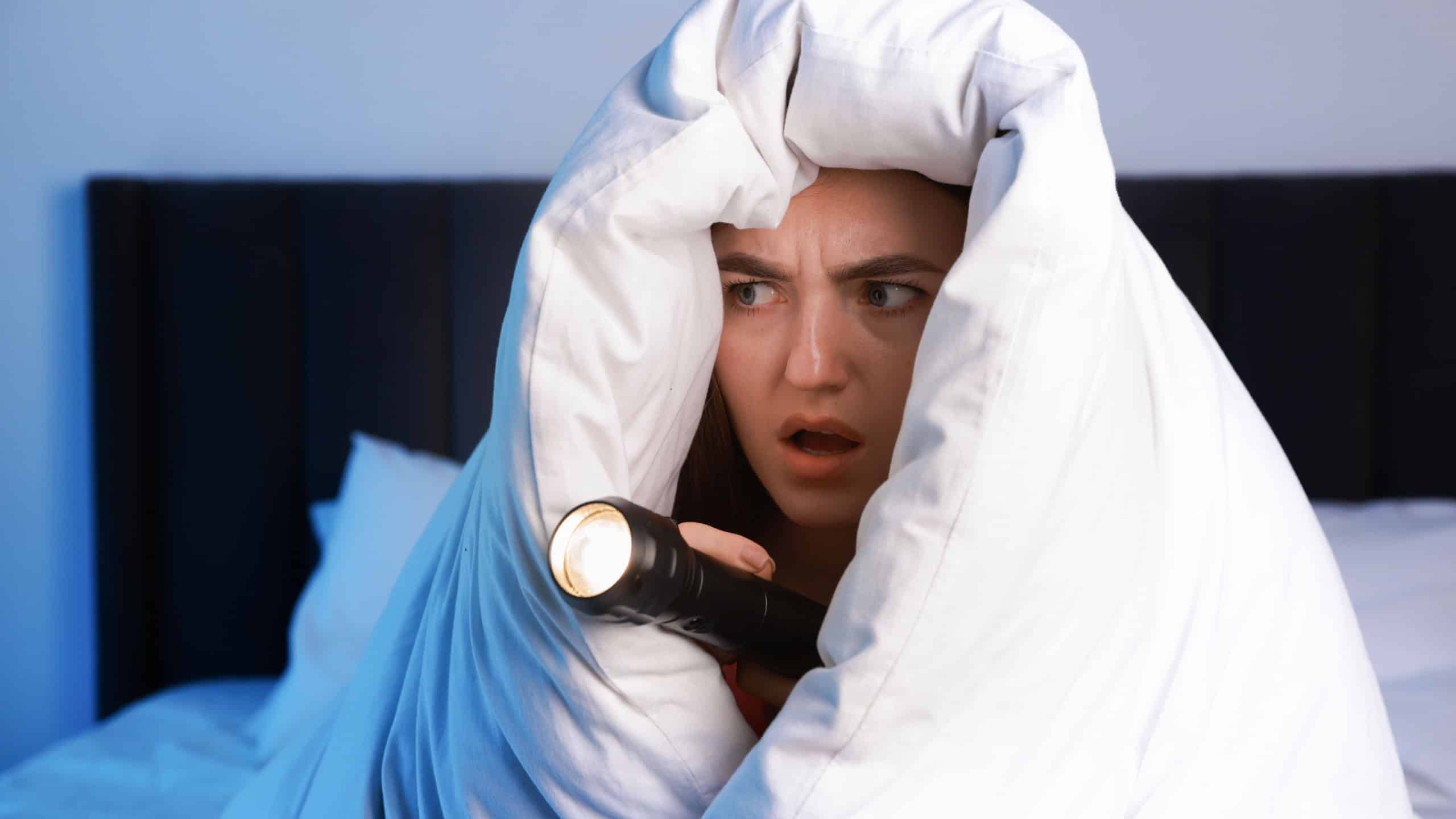 Fear of darkness. Scared young with blanket and flashlight on bed indoors at night