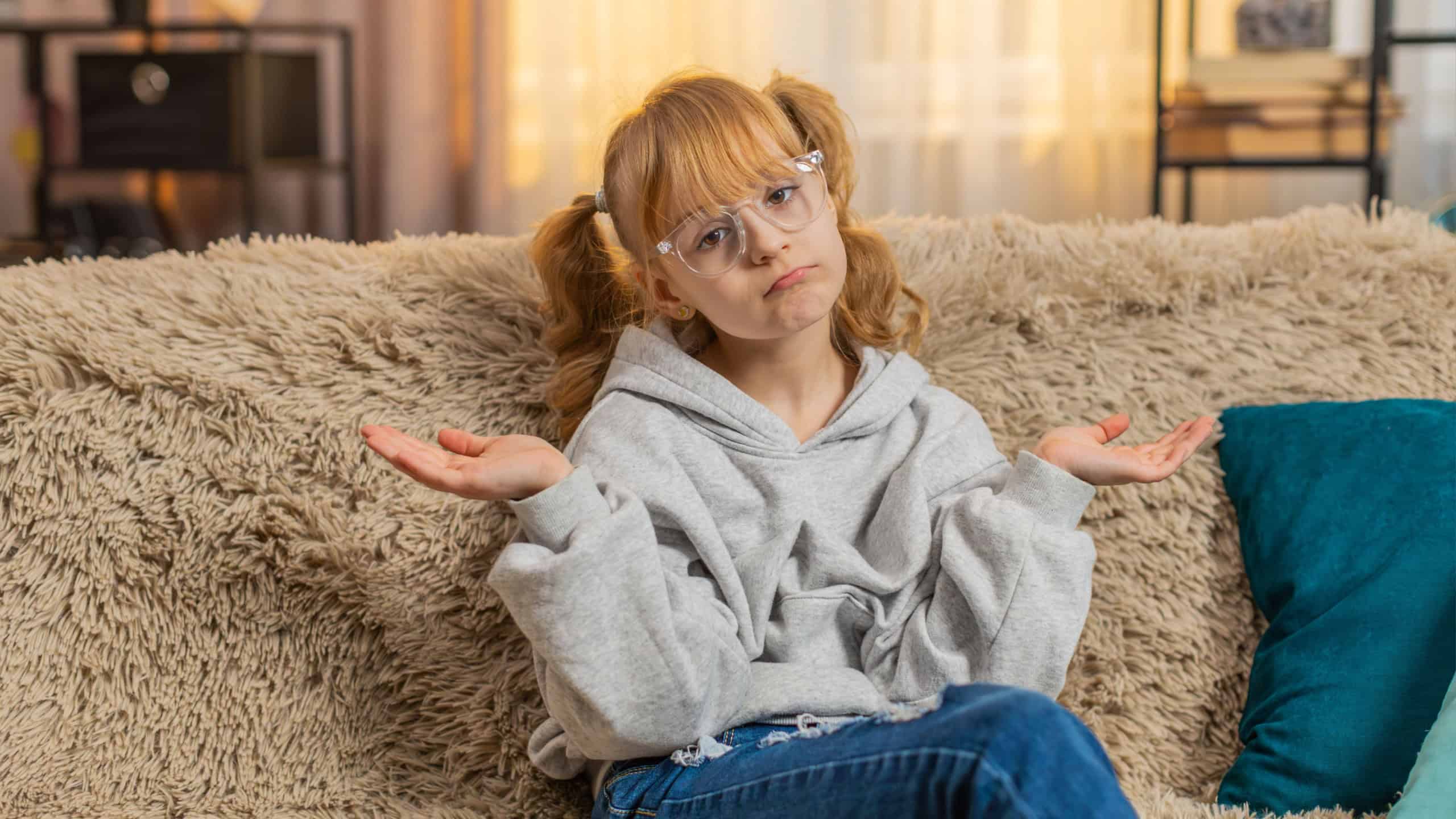 I dont know what to say. Young teenager girl shrugging shoulders with puzzled unsure expression at home. Caucasian child kid sitting on a sofa, showing confusion and uncertainty with relatable gesture