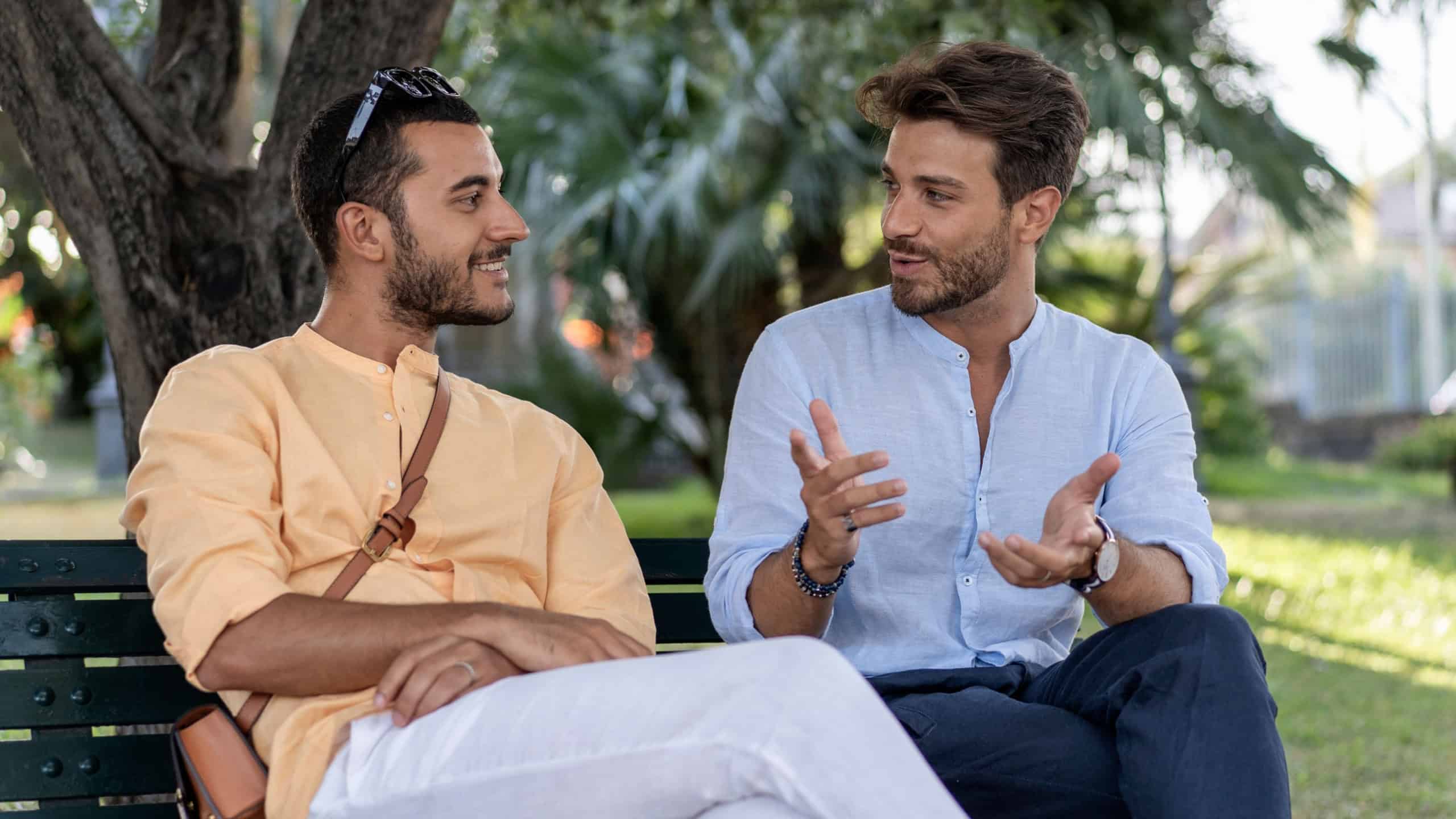 Two young male friends or business colleagues having an engaging and positive conversation while sitting on a bench in a city park. A concept of communication, friendship, and modern lifestyle.