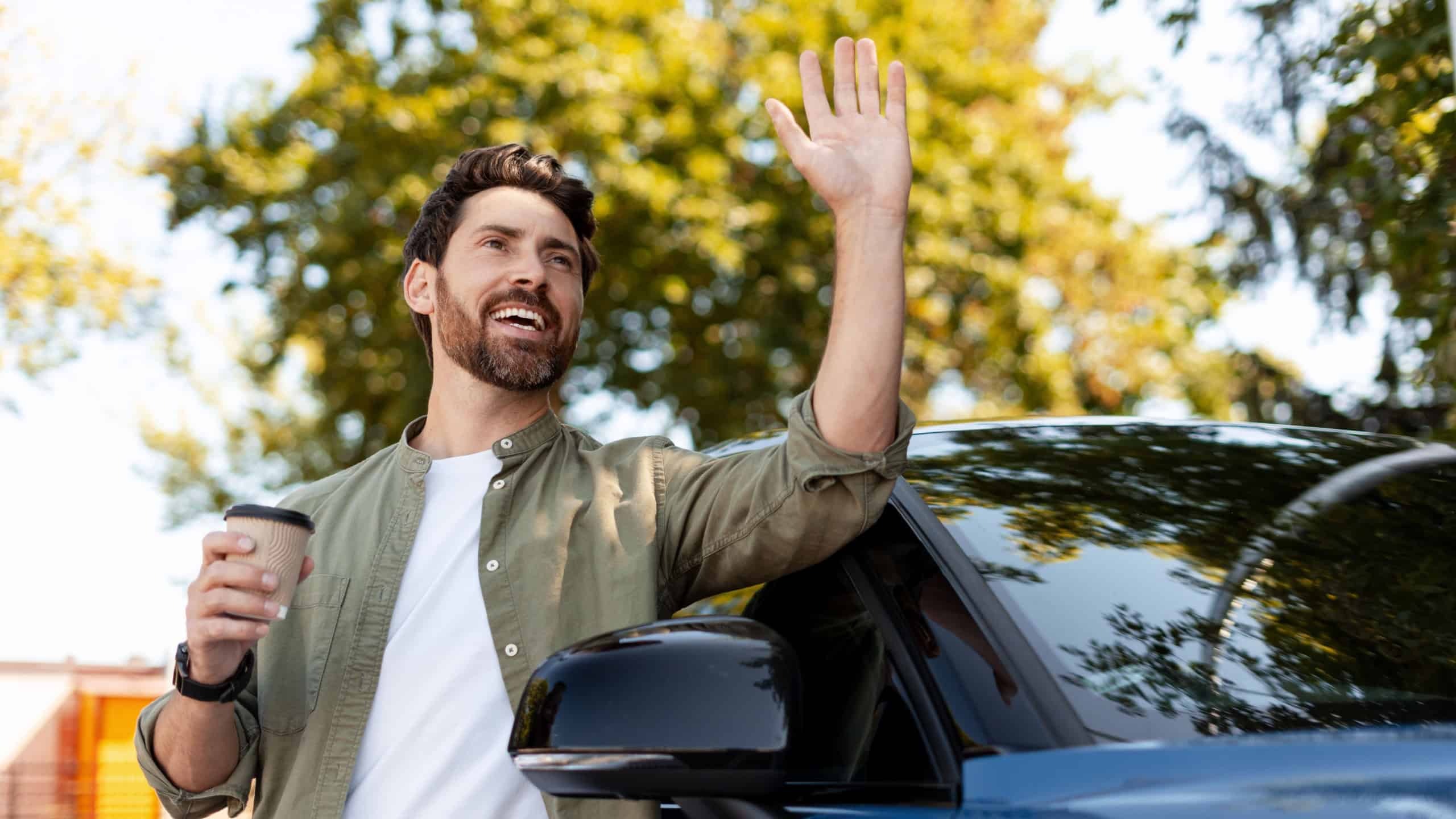 Smiling businessman waving goodbye while holding a coffee near his blue electric car parked outdoors