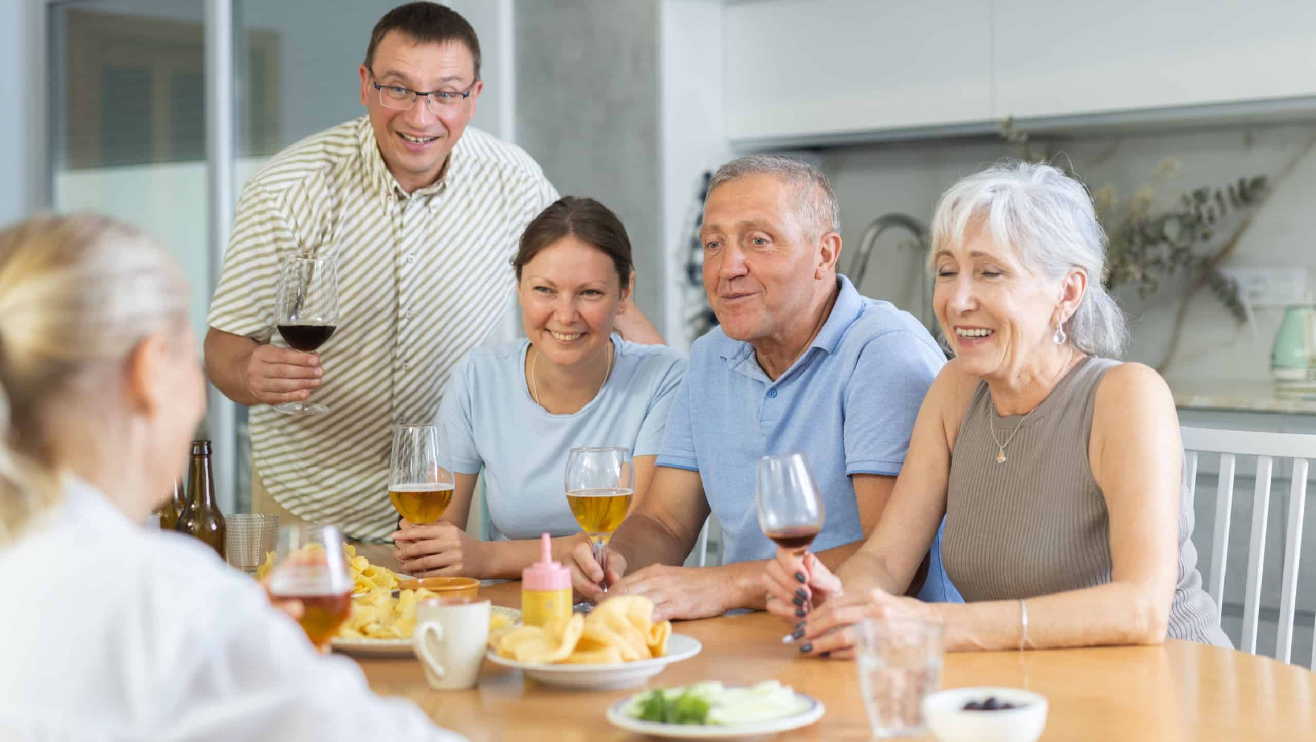 Happy senior couple, man and woman, with adult son and daughter gathering with family friends at cozy kitchen table, chatting cheerfully over drinks with snacks
