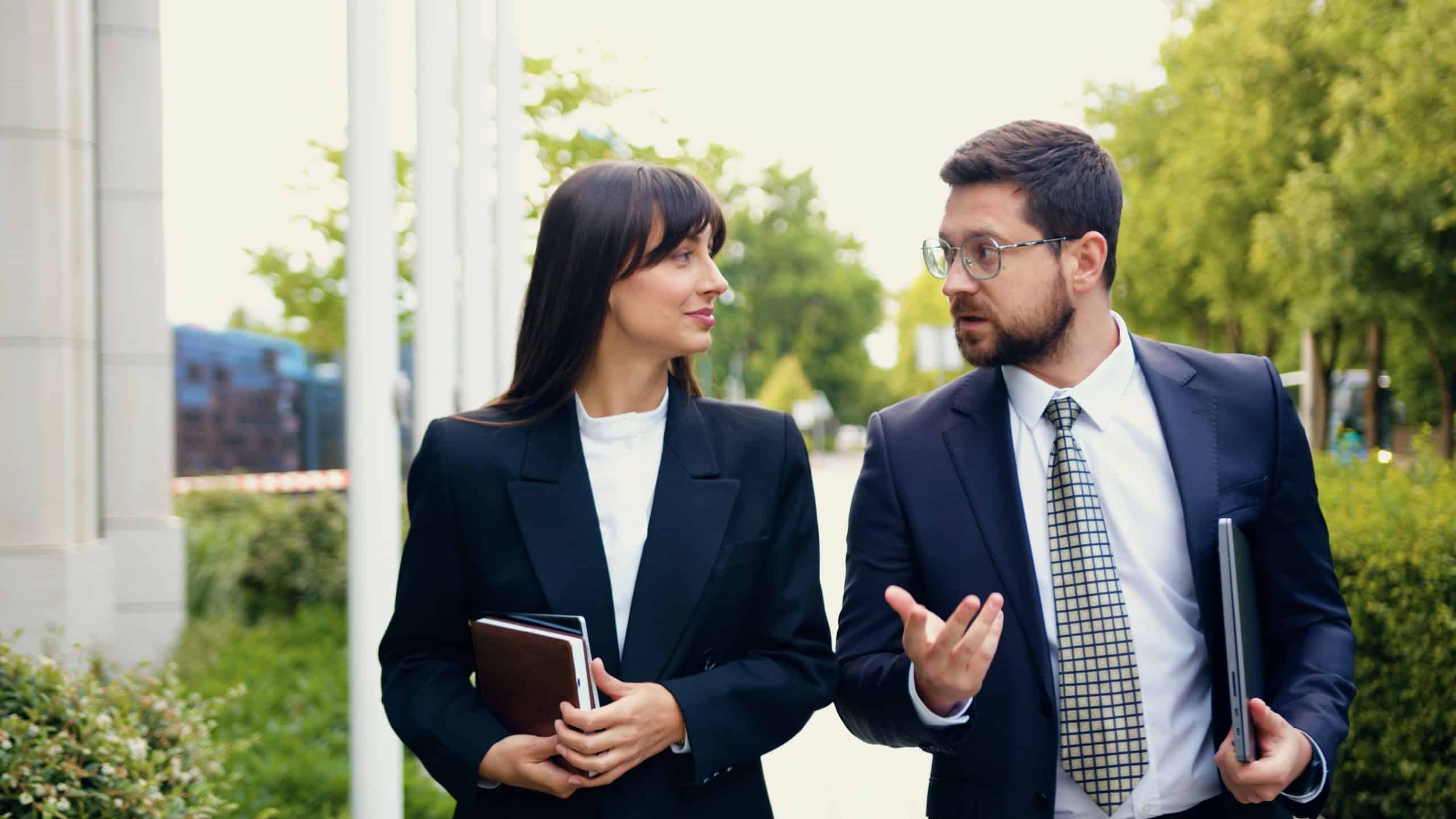 Happy business colleagues, a man and a woman in formal suits, are walking side by side through an urban area, engaged in conversation on their way to work, with the woman carrying a laptop