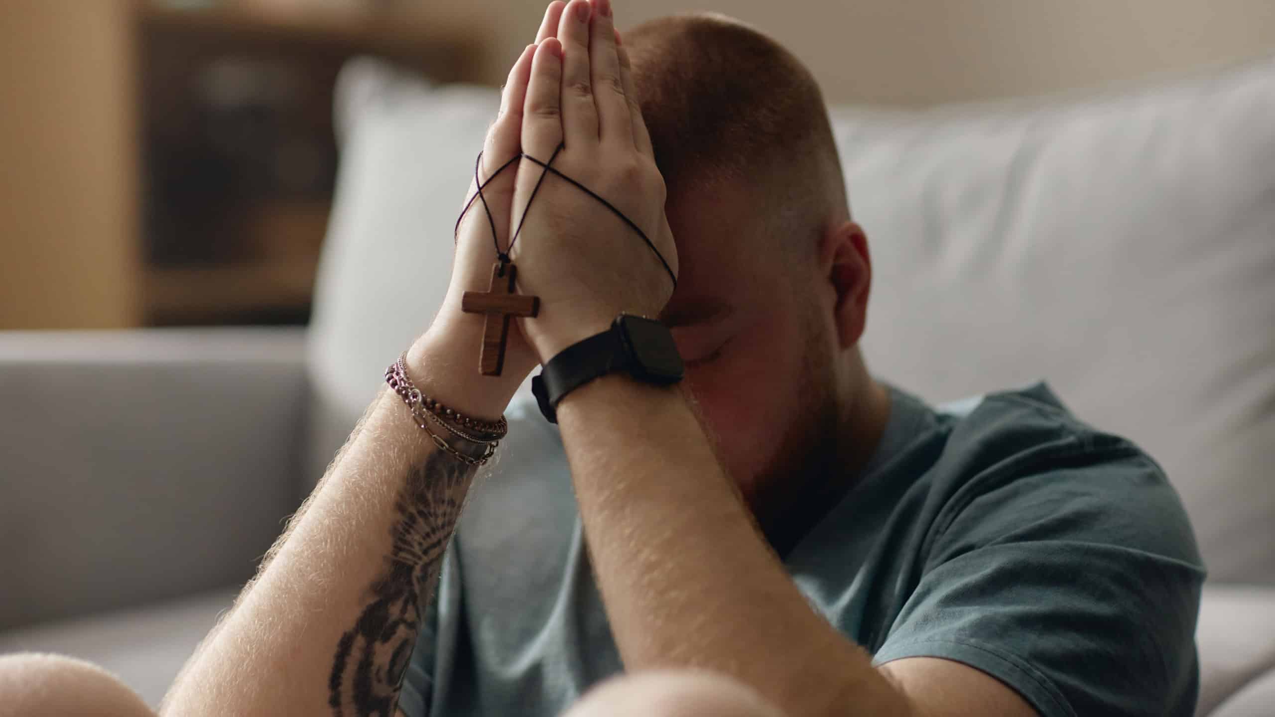 Close up of young adult Caucasian man praying with cross necklace at home, expressing faith, repentance, spiritual struggle. Useful for religion, crisis support, recovery campaigns