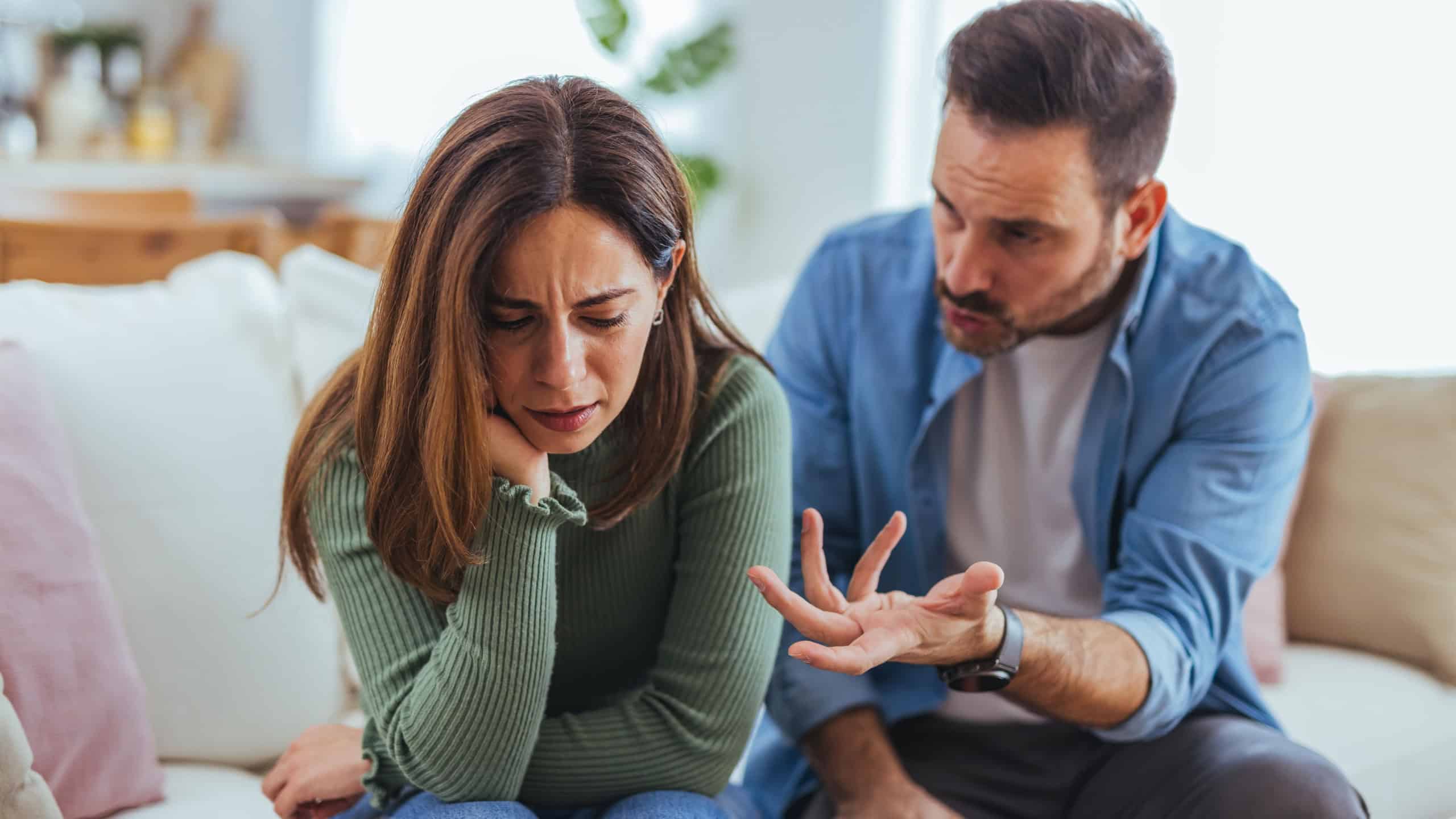 An upset woman sits on a living room couch while her partner gestures and argues, showing emotional tension, frustration and communication breakdown in a domestic relationship scene.