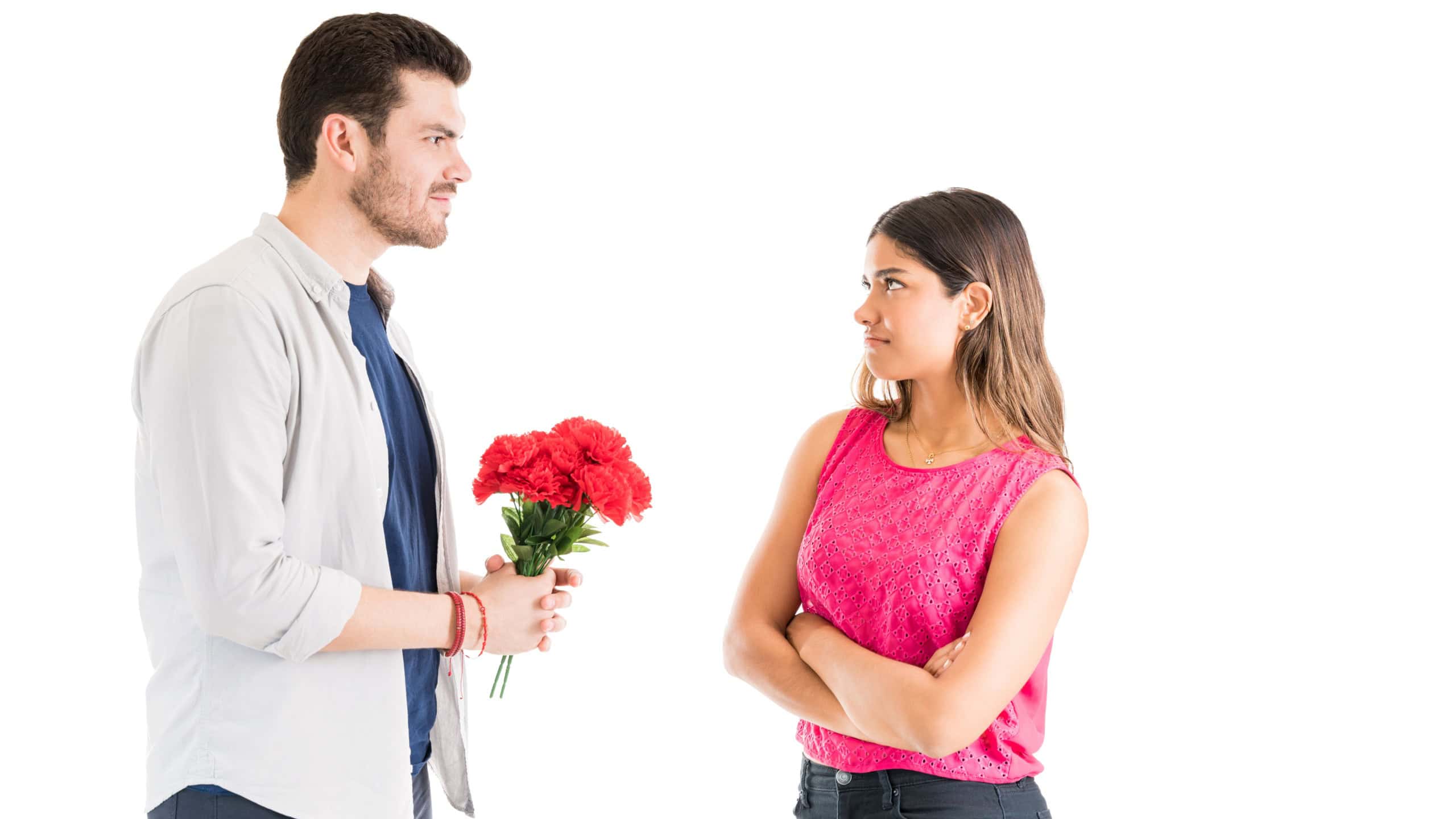 Unimpressed young woman with arms crossed looking at boyfriend holding flower bouquet while standing against white background