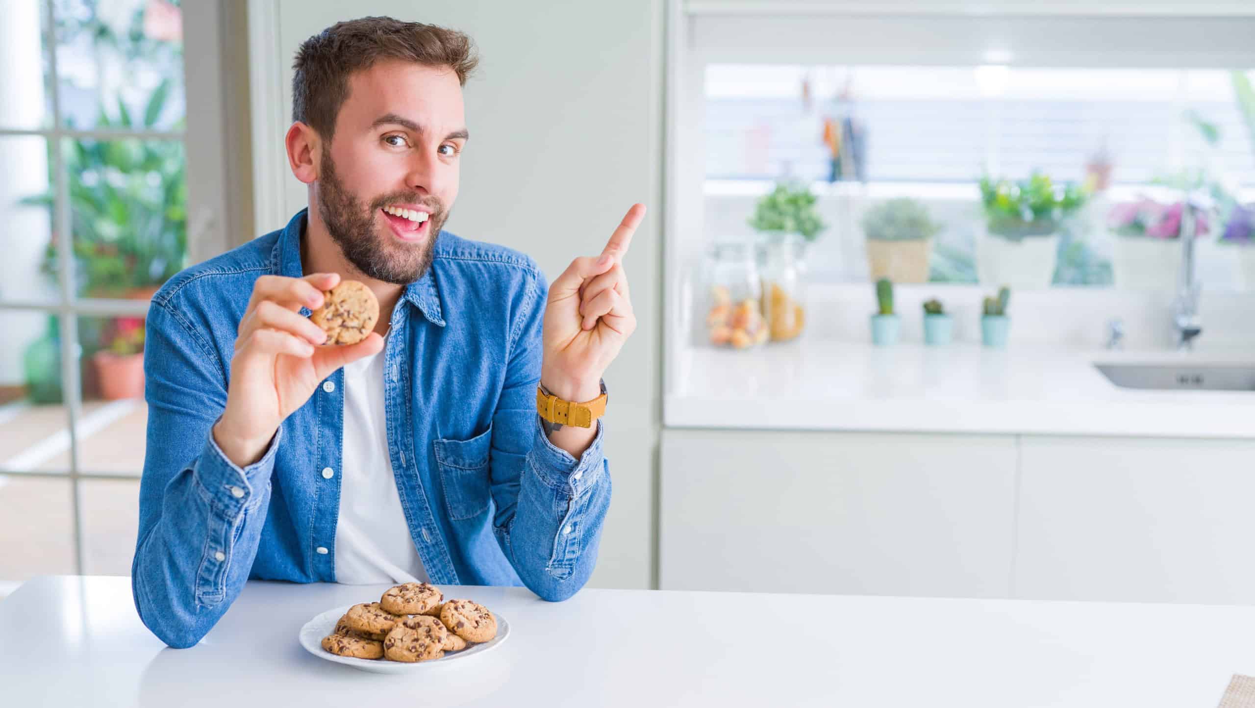 Handsome man eating chocolate chips cookies very happy pointing with hand and finger to the side