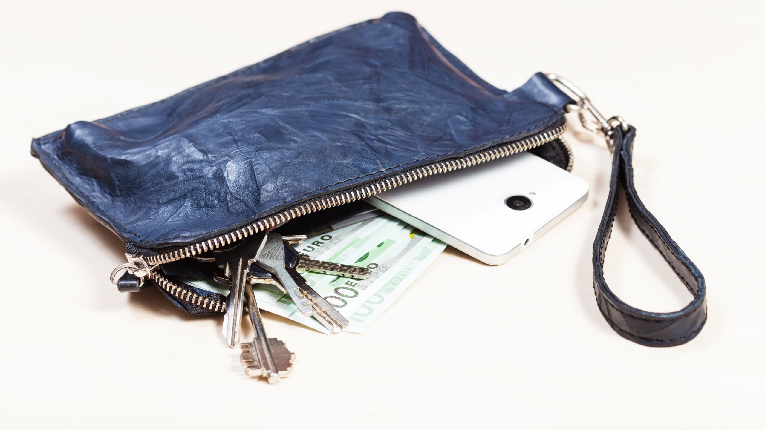 Open blue leather wristlet purse bag with bunch of keys, smartphone and euro banknotes on pale brown table