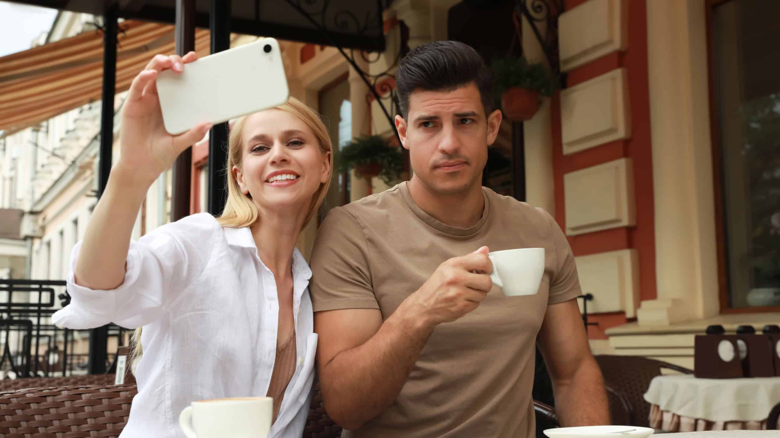 Young woman trying to take selfie with her displeased boyfriend in outdoor cafe. Boring date