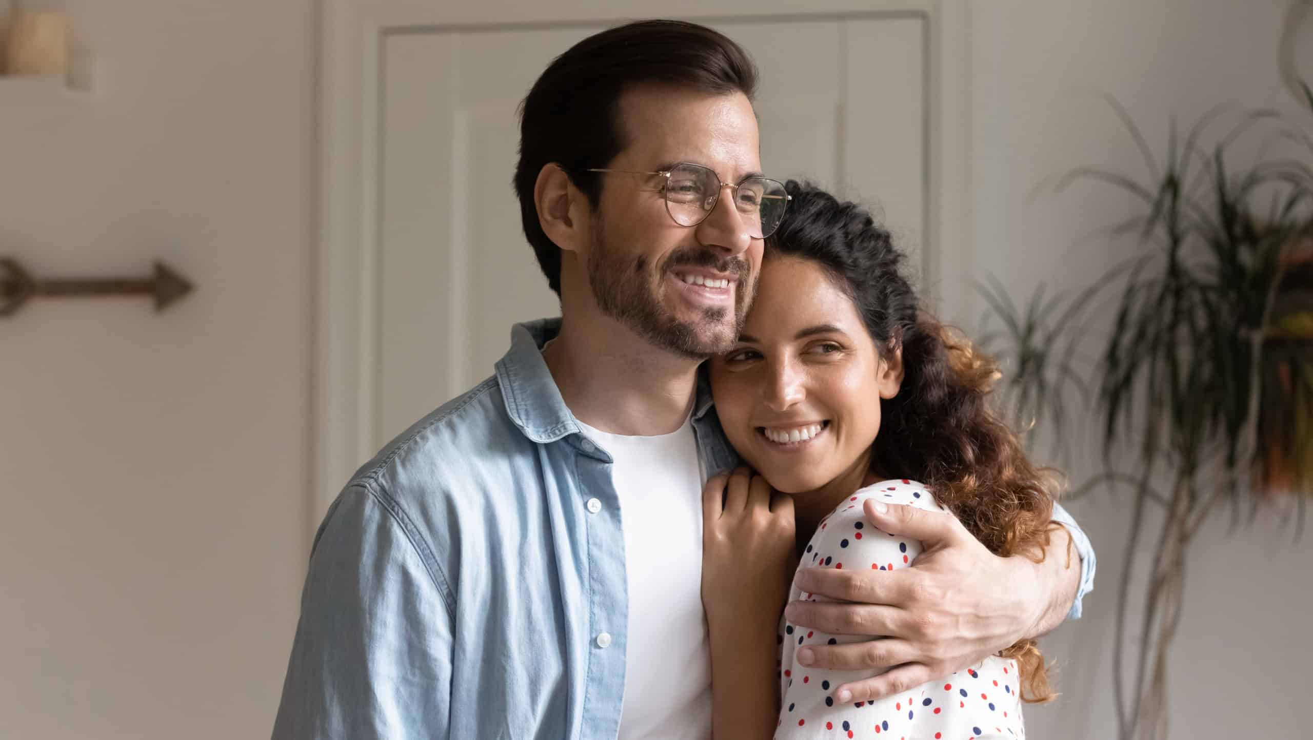 Dreamy 30s couple standing indoor hugging looking out window daydreaming. Husband and wife first time home buyers homeowners feels happy, indulge in dreams about bright future life together concept
