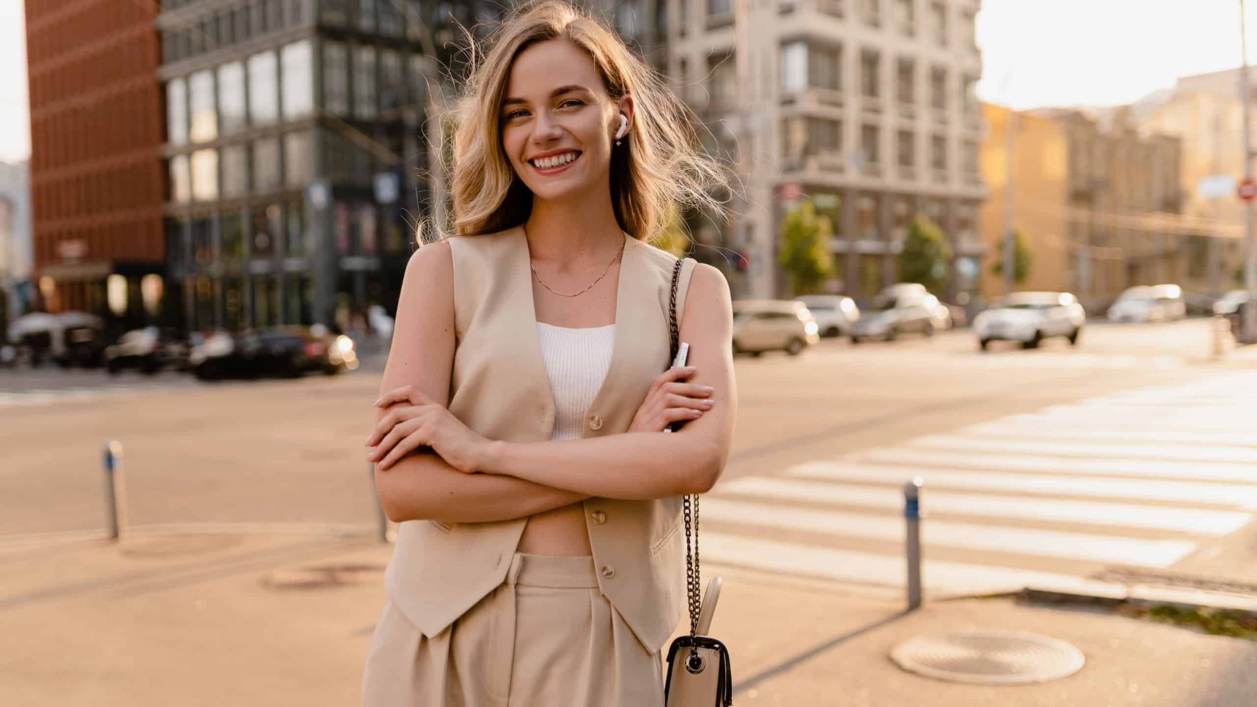 pretty blonde woman walking in sunny city street in beige suit with a purse in urban fashion style, smiling, summer trend season, wearing vest and trousers, glam hair hairstyle